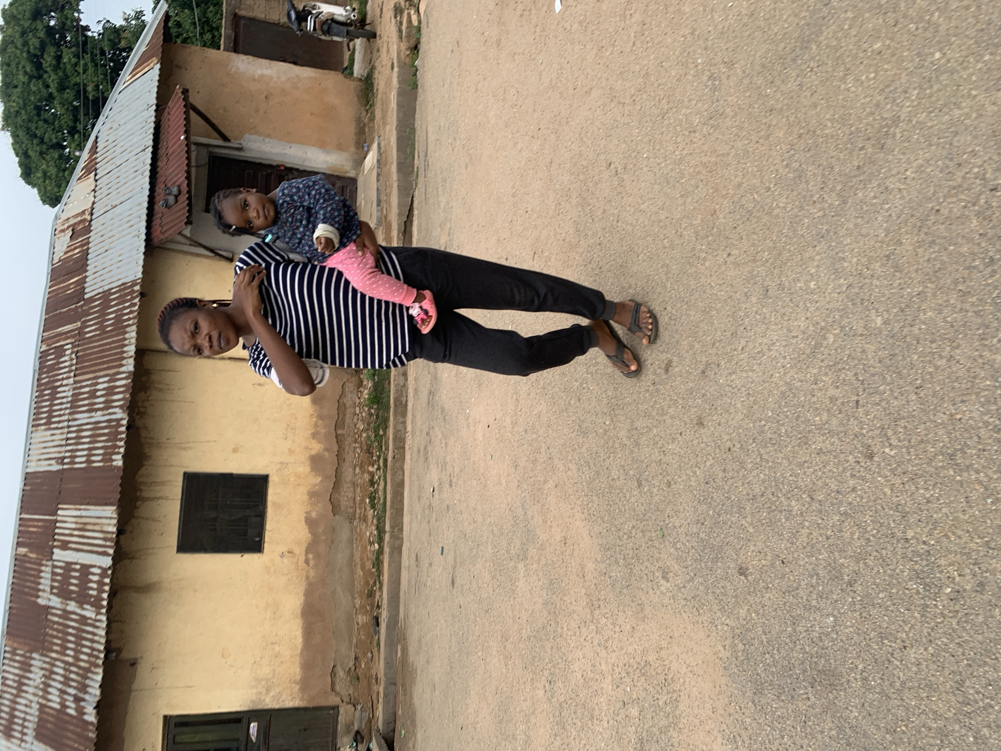 A woman is standing on an asphalt street in Jos, Nigeria, holding a child. The woman wears black pants, a black and white striped shirt, and sandals. The child wears a blue patterned shirt and pink pants. Behind them is a single-story building with a corrugated metal roof, a window, and a door. In the background, a motorcycle is parked. A tree with green foliage is visible in the upper left corner.