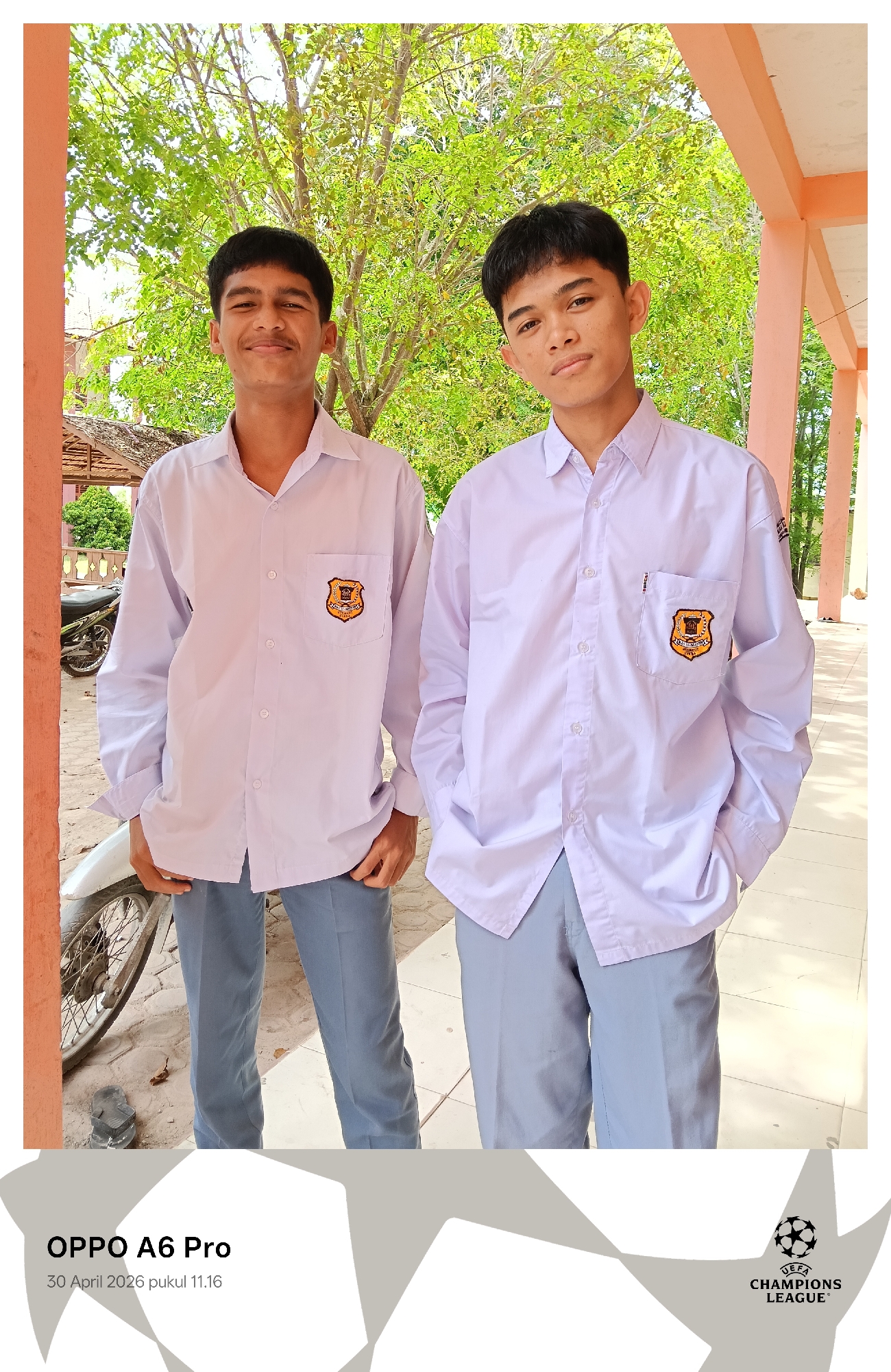 Two young men stand side-by-side outdoors. Both wear matching white shirts with a shield-like emblem on the pocket and light gray trousers. The man on the left smiles, while the man on the right has a more neutral expression. The background features trees, a building structure with orange pillars, and a motorcycle partially visible on the left. The image includes the text "OPPO A6 Pro" and "30 April 2026 pukul 11.16" at the bottom left, and the UEFA Champions League logo at the bottom right. The location is Lhokseumawe, Indonesia.