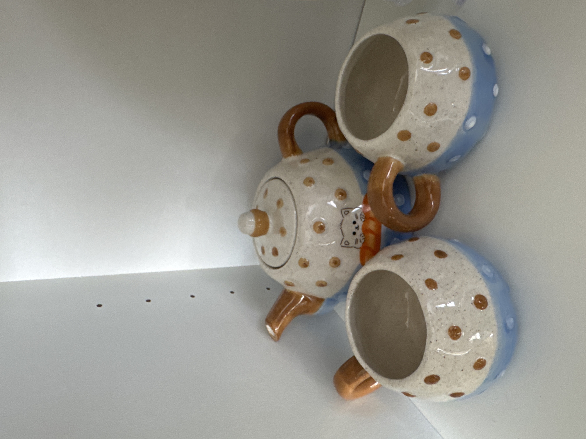 A polka dot teapot and two matching polka dot cups, stacked, are in an enclosed white space, possibly a cabinet. The teapot has a spout and a handle, with a lid on top. A small depiction of a bear is visible on its side. The cups are stacked one on top of the other, with their handles touching the teapot's handle. The predominant colors are white, brown, and light blue. The location of Boreti, Montenegro, is not discernible from the image.