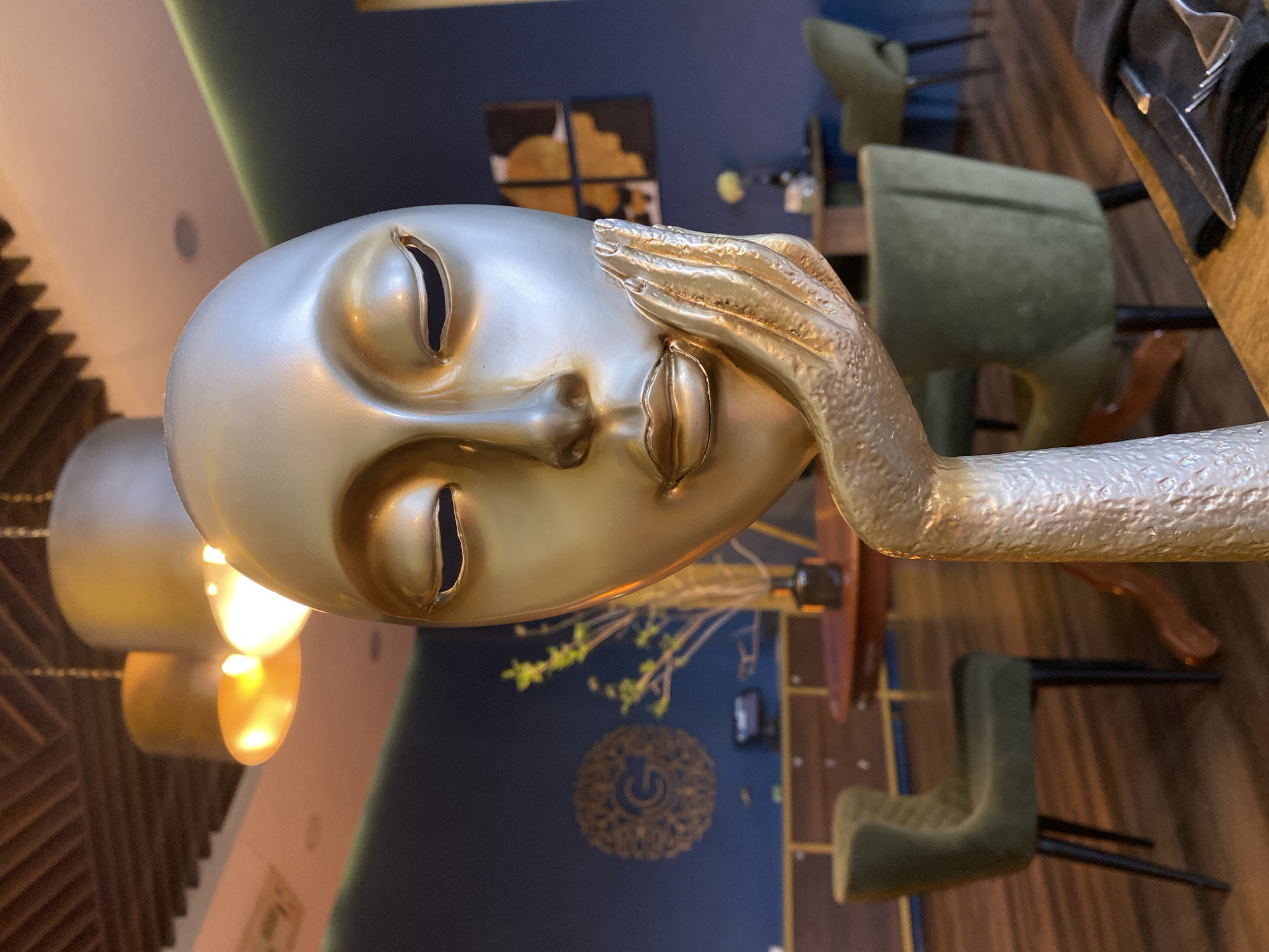 A golden-hued, surreal sculpture of a face with a hand resting on its forehead dominates the image. The sculpture has minimalist facial features, including elongated, slit-like openings for eyes and lips. The background features a deep blue wall adorned with floral decorations and a circular emblem. To the right of the sculpture, there are chairs with green upholstery and a wooden table set with silverware and a dark napkin. A lamp with a multi-tiered, cylindrical shade is visible to the left. The photograph depicts a scene from a restaurant in Yekaterinburg, Russia.