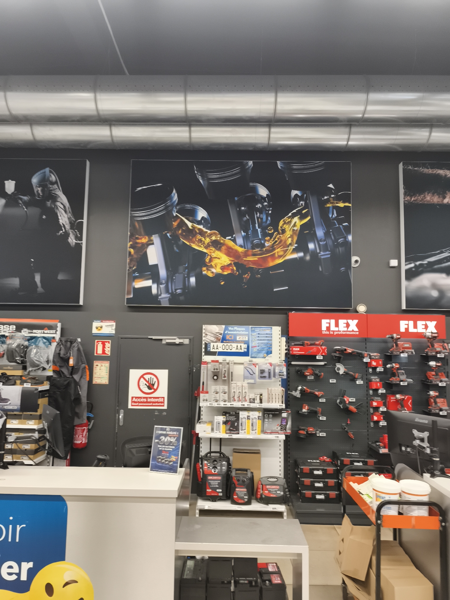 The image shows the interior of a store, likely a hardware or automotive supply store, in Saint-Denis, France. The foreground features a white counter with a blue graphic on the left side and a yellow smiley face icon. Behind the counter, a dark gray door is visible with a red and white "Accès interdit" (Access forbidden) sign. To the right of the door are shelves stocked with red and black power tools, many with the "FLEX" brand prominently displayed. Above the shelves, large framed graphics depicting engine parts and oil are mounted on the dark gray wall. To the left of the door, a section of shelving displays various items including grey workwear and boxed goods. The ceiling features exposed metal ductwork. The overall lighting suggests an indoor retail environment.
