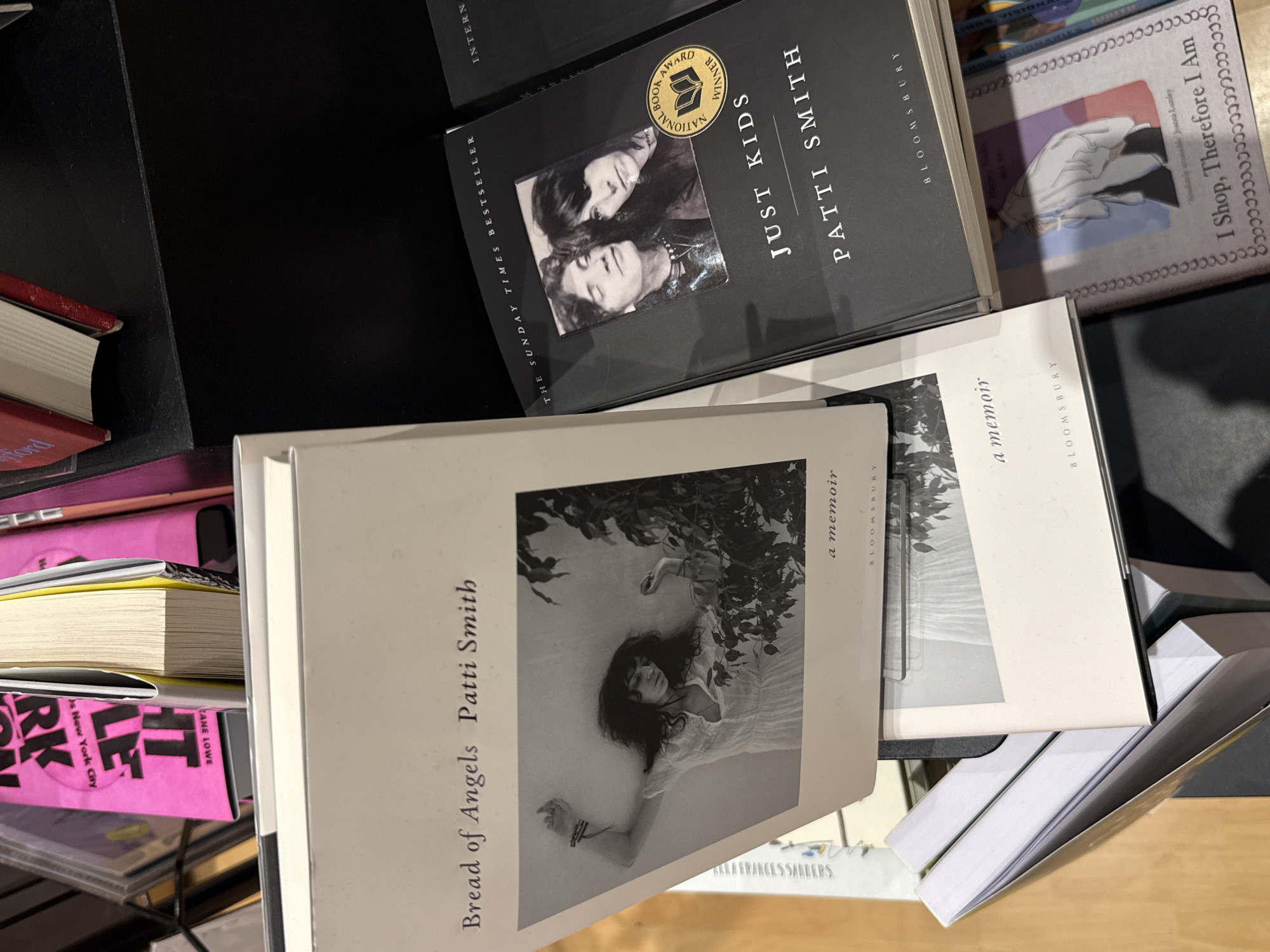 This image shows a close-up of books on a shelf. The books are titled "Just Kids" and "Bread of Angels" by Patti Smith, and "I Shop, Therefore I Am" by Joanna Lumley. The location is not discernible.