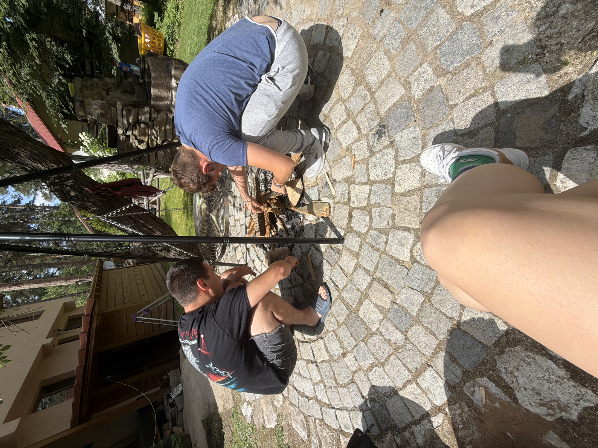 Two men are squatting by a fire pit in an outdoor setting. One man on the left is wearing a black t-shirt, grey shorts, and flip-flops, and is tending to the fire. The man on the right is wearing a blue t-shirt, grey sweatpants, and athletic shoes, and is also interacting with the fire. The fire pit appears to be made of stone. In the background, there is a building with a wooden exterior and a red roof, surrounded by trees and greenery. The ground around the fire pit is paved with cobblestones. The perspective of the image suggests it was taken from a seated position, with a person's legs and feet in the foreground.