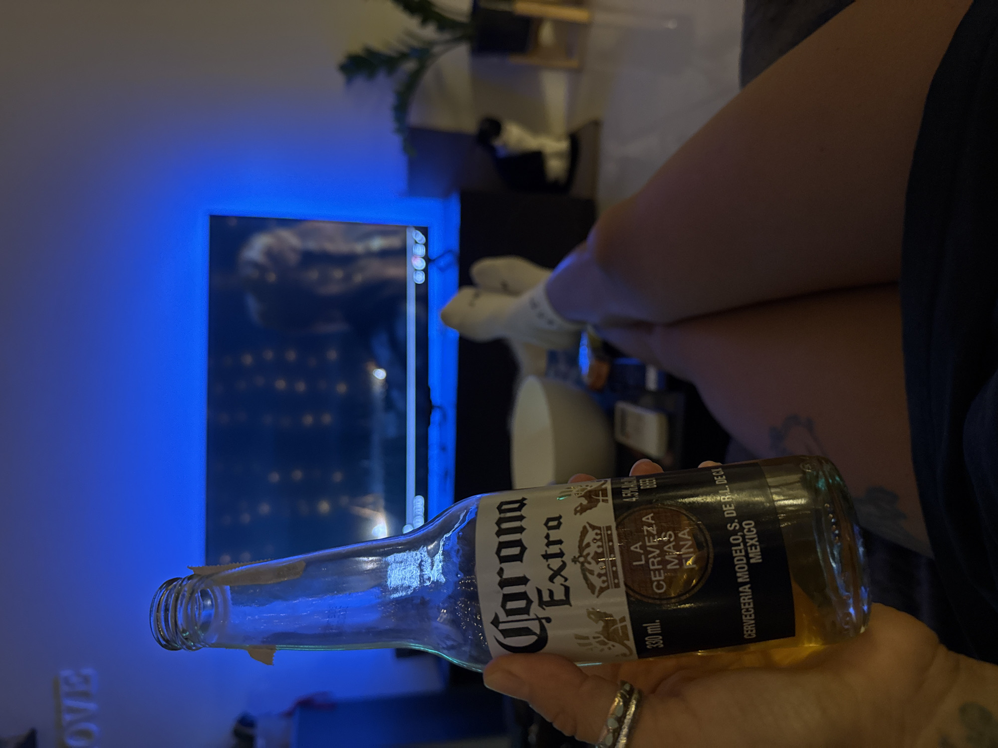A person is holding a bottle of Corona Extra beer, which is partially filled. The background shows a television screen displaying a bright blue light and some abstract shapes. To the right of the TV, a person's legs are visible, wearing white socks. The setting appears to be indoors, possibly a living room, with soft lighting. The overall mood is relaxed and casual. The visible text on the bottle reads "Corona Extra," "LA CERVEZA MAS FINA," "CERVECERIA MODELO, S. DE R.L. DE C.V.," "MEXICO," and "330 ml." The time of day cannot be determined from the image.