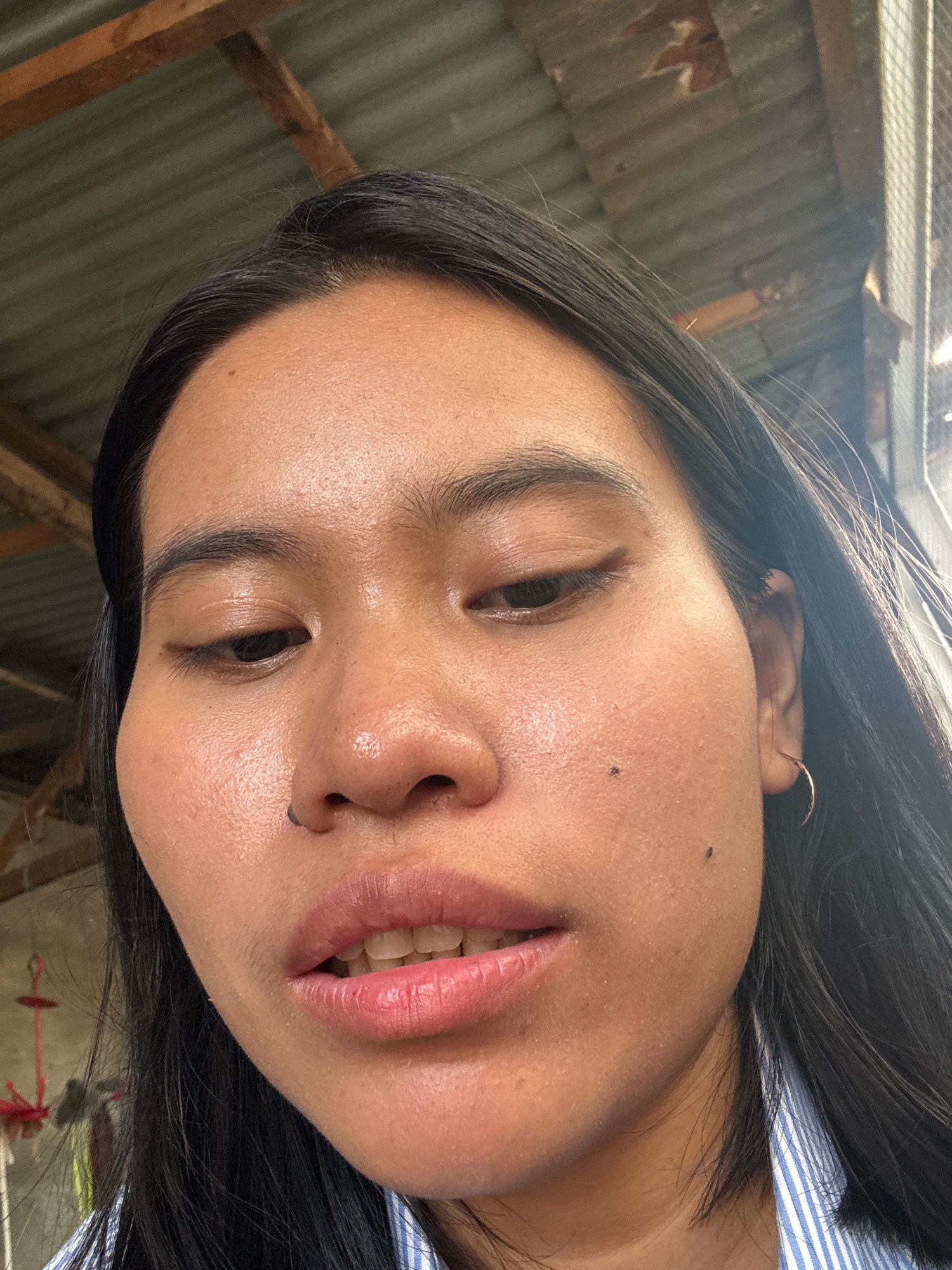 A close-up, low-angle selfie of a young woman's face is captured outdoors. She has dark hair, a medium skin tone, and is wearing subtle eye makeup and lip gloss. Two small moles are visible on her right cheek, and a stud pierces her left nostril. She is wearing a blue and white striped collared shirt. The background consists of a corrugated metal roof structure with wooden beams, suggesting a rustic or outdoor setting, possibly a residential area in East Jakarta, Indonesia. A piece of red material on a string hangs in the far left background. The lighting is natural and bright, indicating it is daytime.
