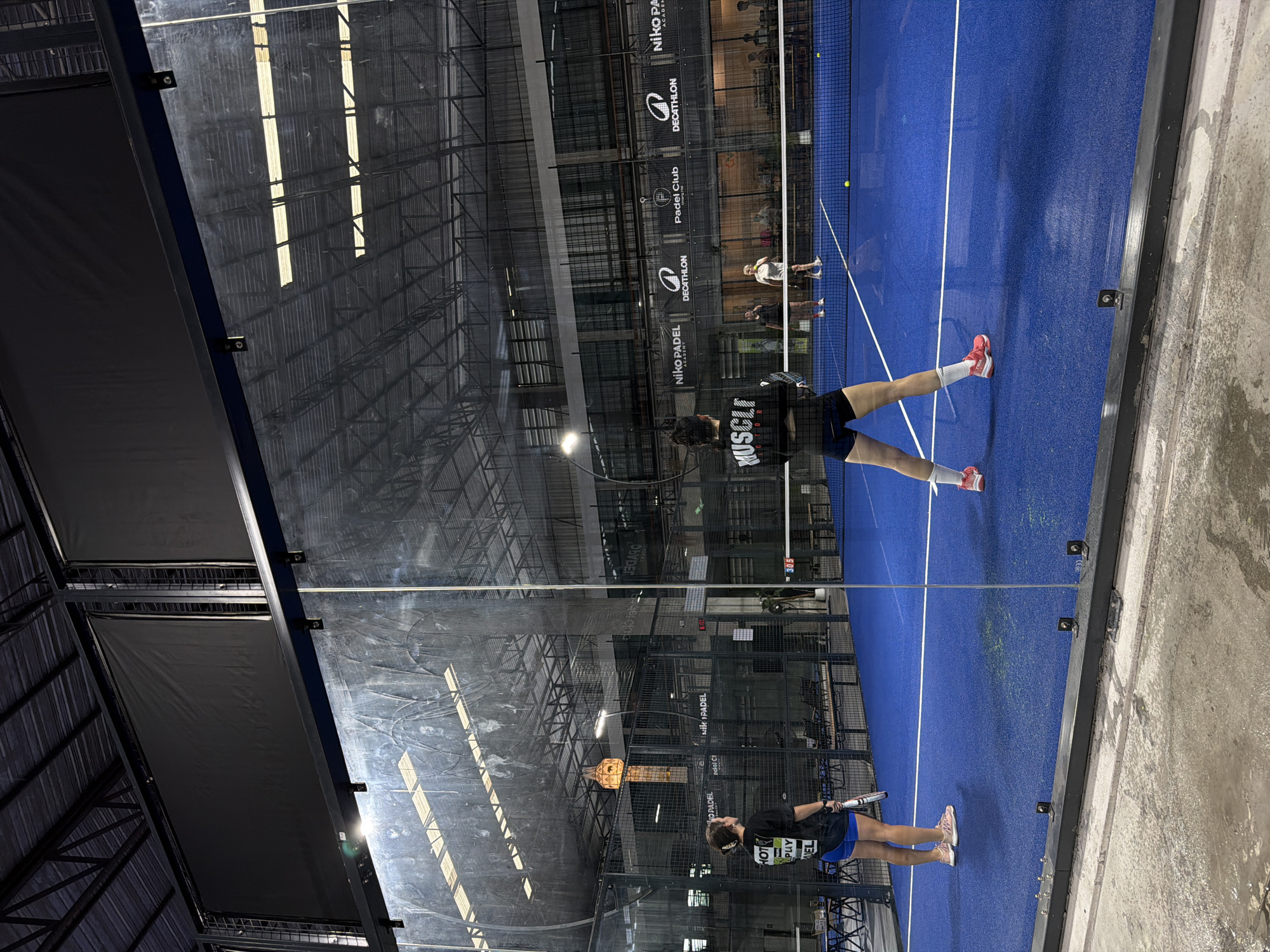 The image depicts an indoor padel tennis court in Ban Buak Khrok North, Thailand. Four individuals are visible on the blue court, engaged in a game. Two women, dressed in athletic wear, are on one side of the net, with one in the foreground facing away from the camera, holding a racket. The other two players are on the opposite side of the net, also participating in the match. The court is enclosed by glass walls and metal fencing, with a netted ceiling above. Banners bearing logos such as "Niko Padel Academy" and "Decathlon" are displayed on the back wall. The lighting suggests an indoor facility, with overhead lights illuminating the playing area. The surrounding environment appears to be a sports complex, with seating areas and other courts possibly present in the background.