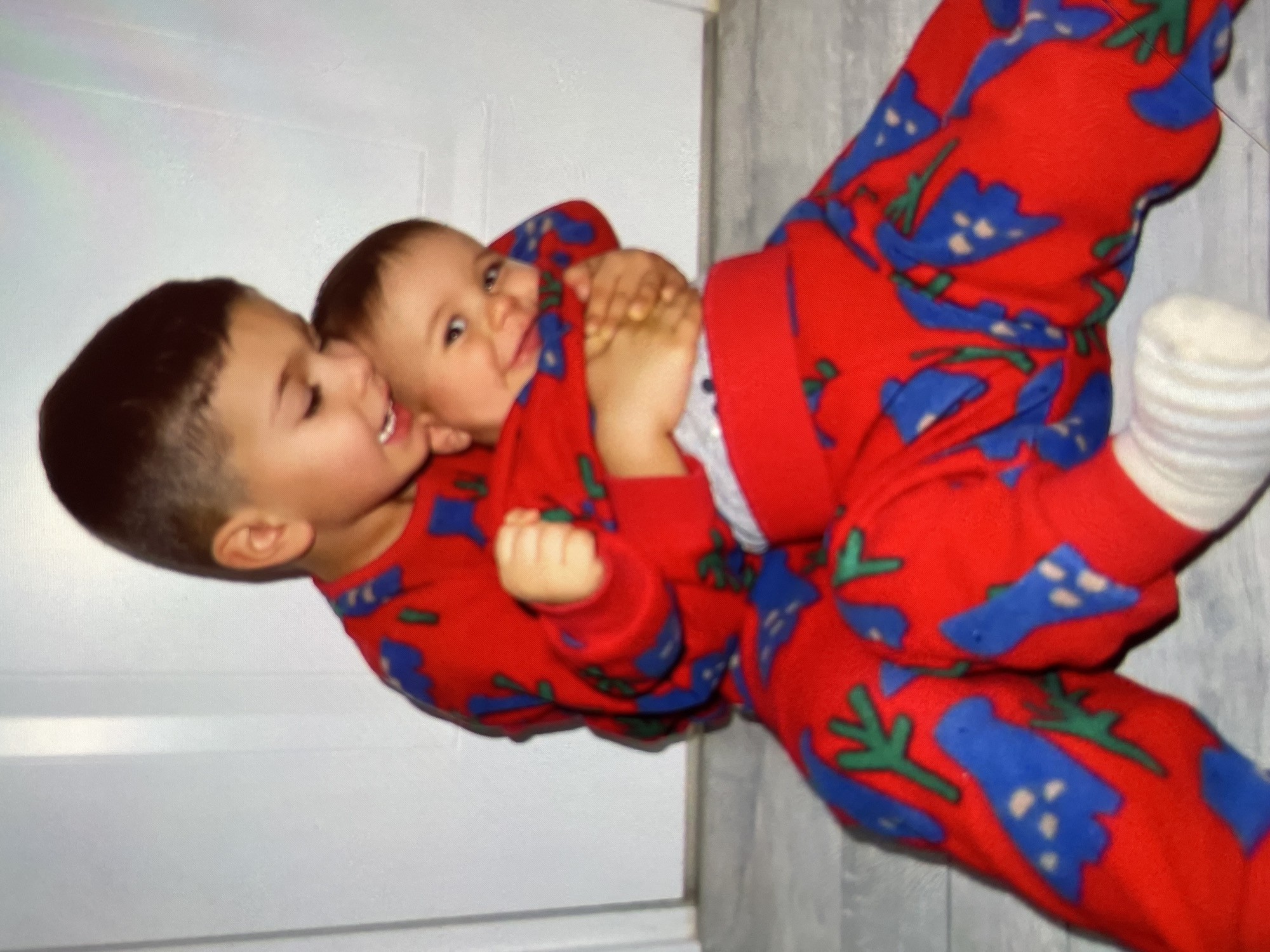 Two young children, both wearing red pajamas with a blue floral pattern, are interacting. One child, seemingly older, is embracing the other, who is lying down with their head towards the right. The older child is smiling, while the younger one is looking at the camera. The background consists of a white wall and a light-colored floor with wood grain detail. The scene appears to be indoors. There is no information to suggest this photo was taken in Harmanli, Bulgaria.