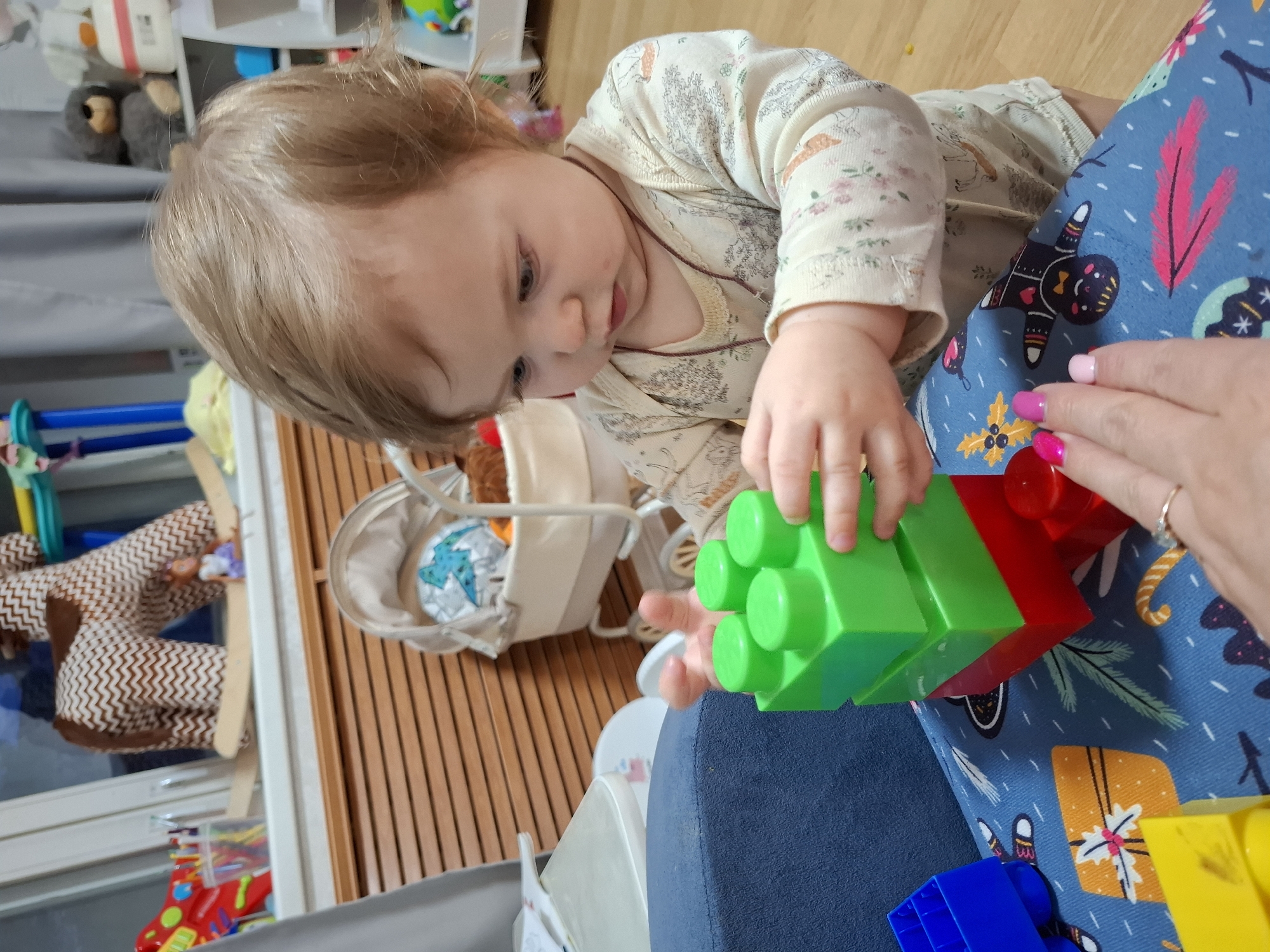 An indoor scene in Chaikovsky, Russia, captures a baby playing with large building blocks. The baby, with light brown hair, is wearing a long-sleeved, light-colored onesie patterned with trees and animals. They are intently focused on two connected blocks, one green and one red, held in their hands. An adult's hand, with pink and fuchsia nail polish and a ring on one finger, is visible near the blocks, suggesting shared play.
The baby is leaning over a surface covered by a patterned blanket or garment, which features festive designs including a gingerbread man, gift boxes, holly, and pine branches on a blue background. Other colorful blocks, including blue and yellow ones, are scattered nearby.
In the background, a wooden slatted floor is visible. Various toys and household items are blurred, including what appears to be a baby bouncer or seat with a white fabric lining and a patterned cushion, and a brown and white zig-zag patterned stuffed animal or rocking toy. The lighting suggests it is daytime. The overall atmosphere is one of calm, focused play.