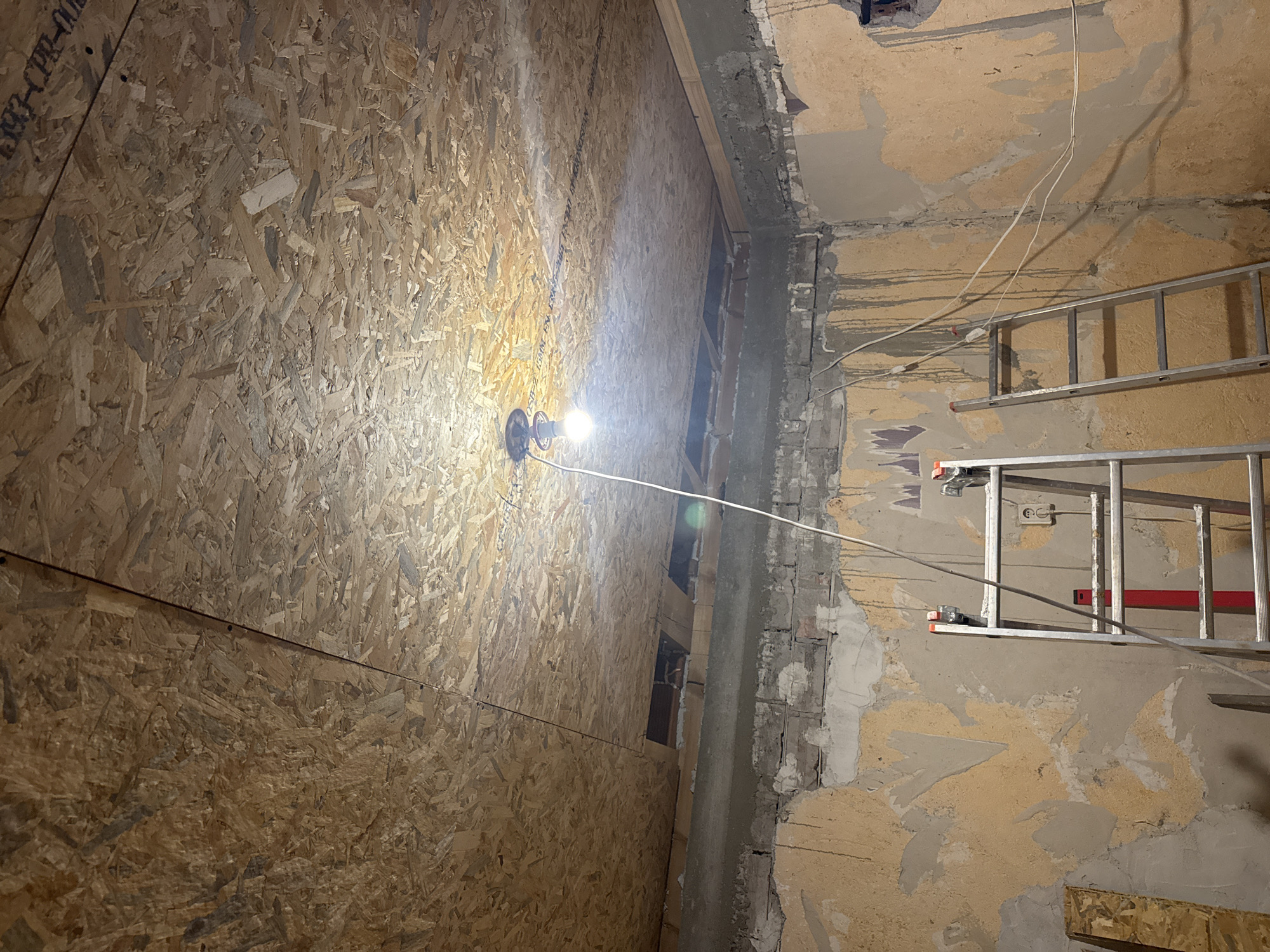 An interior scene within a building, stated to be located in Sarnevo, Bulgaria, depicts a room undergoing renovation or construction. The ceiling is predominantly covered with Oriented Strand Board (OSB) panels, fastened to an unseen frame, with a visible seam running diagonally from the upper right toward the center-left. Printed text, including "OSB/2 EN 300-2006" and "1393-CPR-001", is partially legible on some panels. A temporary, bare light bulb, suspended by a white electrical cord from a black fixture, hangs centrally from the OSB ceiling, providing the primary illumination. Near the back-left and back-right sections of the ceiling, rectangular cutouts reveal the structural elements above, including exposed brickwork and support beams.
Below the ceiling, a rough concrete beam or lintel spans the upper perimeter of the room. The walls below this beam are in a state of disrepair, exhibiting multiple layers of exposed material. Patches of faded beige or yellowish old plaster or paint are visible, alongside areas where these layers have peeled away, revealing gray concrete and sections of underlying brickwork. Irregular dark vertical streaks, possibly indicative of moisture or old adhesive residue, are present on the right wall. Exposed white electrical wiring runs along the upper part of the right wall, with a single two-pin electrical outlet (Schuko type) partially visible on the same wall. A rough, dark opening is present in the upper right corner of the right wall.
In the foreground, two silver-colored, multi-step ladders are present. The more prominent ladder, positioned centrally, is an A-frame step ladder with red stabilizing accents. A second ladder, possibly an extension ladder or another step ladder, leans against the right wall, partially obscured. A rectangular piece of OSB or unfinished wood leans against the lower section of the left wall. No individuals are present in the scene, which suggests active work is either paused or pending.