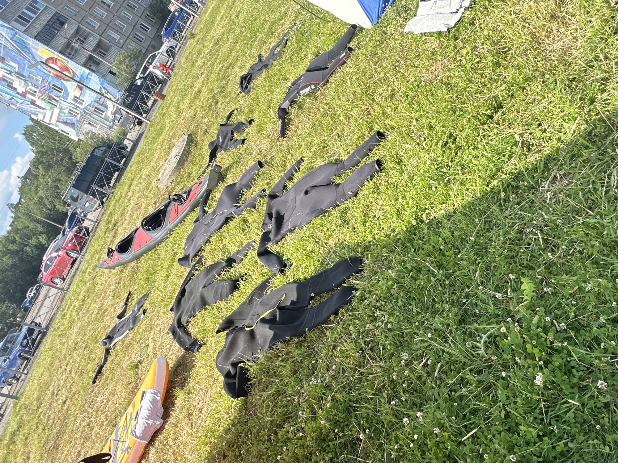 The image depicts a grassy area outdoors, possibly a park or a yard, in the city of Monchegorsk, Russia. A variety of wetsuits are spread out on the grass, suggesting preparation for or aftermath of water activities such as kayaking or paddleboarding. One wetsuit is predominantly black with some yellow detailing. Other wetsuits are also black. A red and grey kayak is visible in the background, along with a yellow and orange paddleboard covered with white material. Beyond the grassy area, a street is lined with parked vehicles, including a blue SUV, a red car, and a black van. In the far background, a multi-story apartment building with a large mural on its side is present. The sky is clear and blue with some clouds. The scene is illuminated by bright daylight, casting shadows on the grass. There are no people visible in the immediate vicinity of the wetsuits and watercraft.