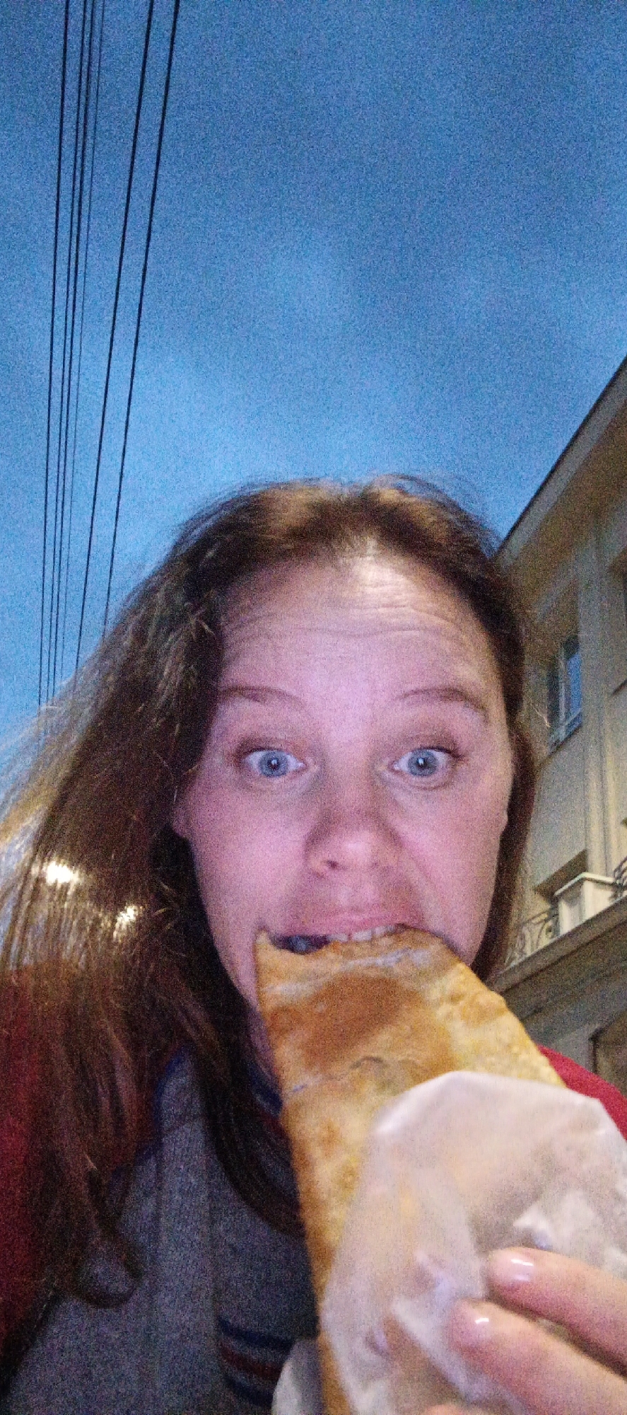 A person is taking a selfie outdoors in Votkinsk, Russia. The sky above is a twilight blue with several dark power lines visible. To the right, a building with windows and balconies is partially visible. The person, seen from the chest up, has long, brown hair and is looking directly at the camera with wide blue eyes. They are in the process of taking a bite out of a large piece of food wrapped in white paper. The food appears to be a savory pastry or pie. The person is wearing a gray sweater with red and blue stripes on the collar and a red jacket. Their left hand is visible, holding the food.