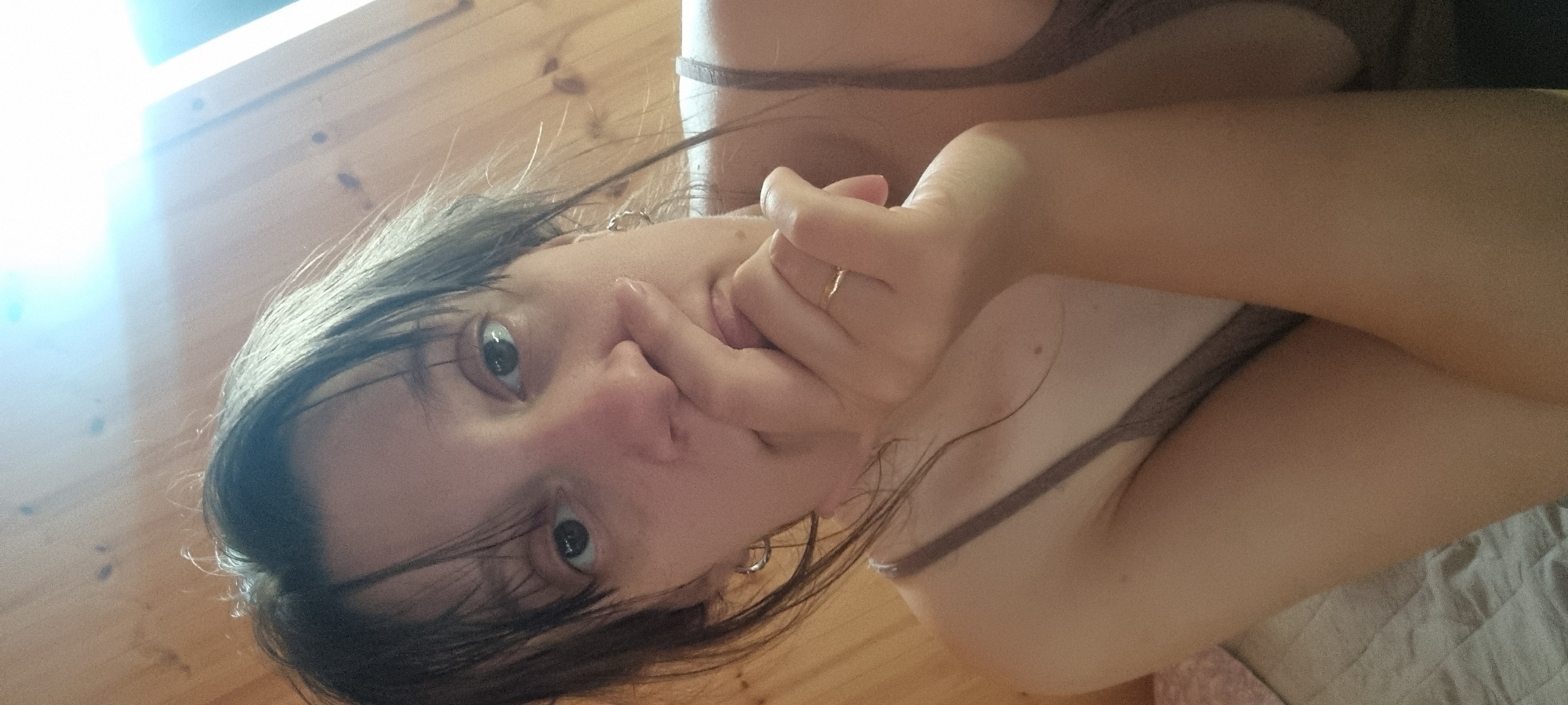 The image shows a close-up, medium shot of a young person, depicted with a 90-degree clockwise tilt. The subject, who has dark hair, is looking directly at the camera with wide, open eyes. Their right hand is raised towards their face, with a finger gently touching their nose or upper lip, creating a thoughtful or slightly shy expression. A thin, gold-colored ring is visible on a finger of this hand, and a silver-colored hoop earring is in their visible ear.
The person is wearing a light brown or tan-colored top with thin straps, parts of which are visible over their shoulders and chest. The background consists of light-colored wooden planks, suggesting an indoor setting with a wooden wall or ceiling. A bright, intense light source causes a significant glare in the upper left portion of the image, indicating daytime. In the lower right quadrant, a textured, light-colored fabric, likely a blanket or bedding, is partially visible. No text is present in the image.