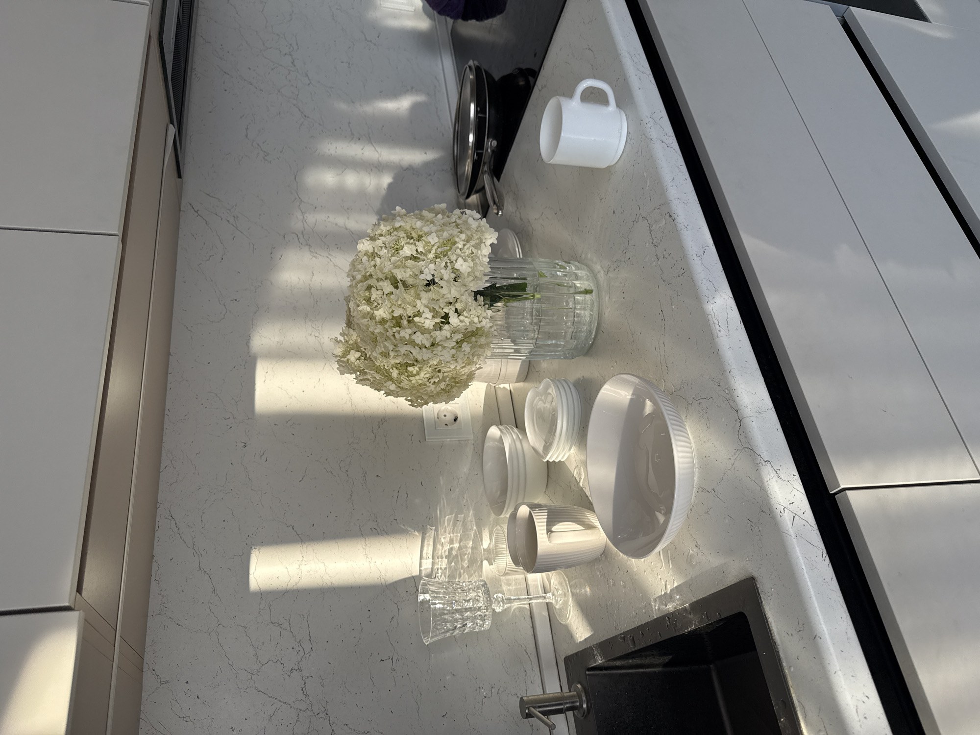 A sunlit kitchen counter in Nizhneyarkeyevo, Russia features a variety of white tableware and a vase of white flowers. On the left, a black sink is visible with a silver faucet. To the right of the sink, a clear crystal wine glass, a ribbed white mug, and several stacked white bowls are arranged. In the center, a large clear glass vase holds a bouquet of white hydrangeas. To the right of the flowers, a small white mug sits on the counter. In the background, a black stovetop with a frying pan is visible. The countertop is white with a subtle gray veining pattern, and light streaks across it from the sun.