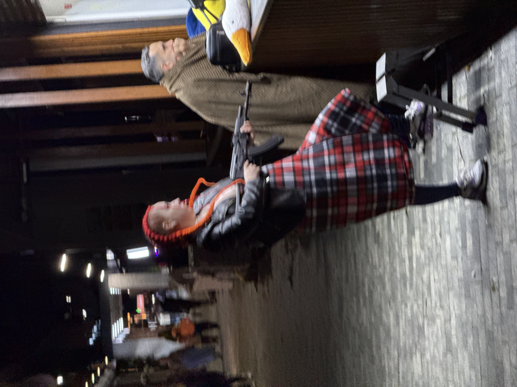 The image appears to be taken at night in an outdoor urban setting. A person wearing a red and orange head covering, a black leather jacket, and a red and black plaid skirt is holding what appears to be an AK-47 style rifle. The person is wearing grey sneakers. Behind them, there is a mannequin wearing a tan coat with a stuffed goose. There are blurred lights and indistinct shapes in the background, indicating other people or buildings. The ground appears to be paved with light-colored, narrow bricks. There is no reliable information in the image to conclude that this is Sochi, Russia.