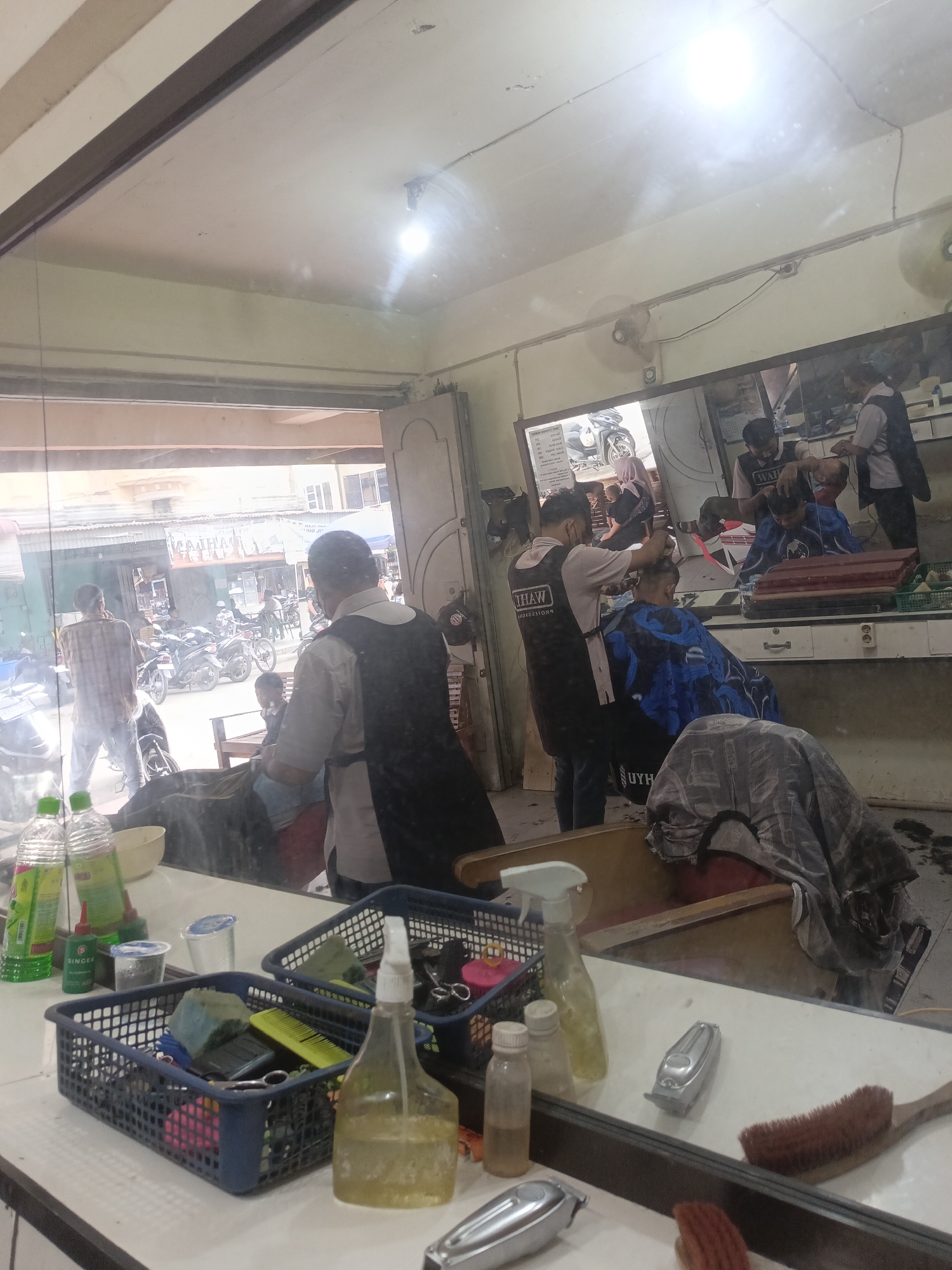 This image depicts the interior of a barbershop in Lhokseumawe, Indonesia, viewed through a mirror. The scene is bustling with activity. Several barbers are attending to customers, with one barber in the foreground using clippers on a customer's head, who is draped in a blue cape. Another barber in the background is also cutting hair. The reflection in the large mirror behind the barbers reveals more individuals and the street outside, showcasing a typical Indonesian urban scene with motorcycles and shops.
The time of day appears to be daytime, as indicated by the bright lighting, possibly from the sun filtering in from the open doorway and the overhead fluorescent lights, which create reflections on the mirror's surface. The weather is not directly discernible but the bright light suggests it is likely fair.
Prominently displayed on the counter in the foreground are various barbering tools and supplies. Two dark blue plastic baskets hold an assortment of items, including combs, scissors, brushes, and spray bottles. A large yellow spray bottle and a smaller clear spray bottle are also visible. A silver electric hair clipper and a brown brush lie on the counter further to the right. In the background, through the open doorway, a street scene is visible with several motorcycles parked.
One of the barbers in the foreground wears a black apron over a light-colored shirt, with the word "TIHAW" visible in white lettering on the apron. Another barber in the background also wears a similar apron. The overall atmosphere appears to be professional and busy, typical of a local barbershop. The image captures a slice of everyday life and commerce in Lhokseumawe.