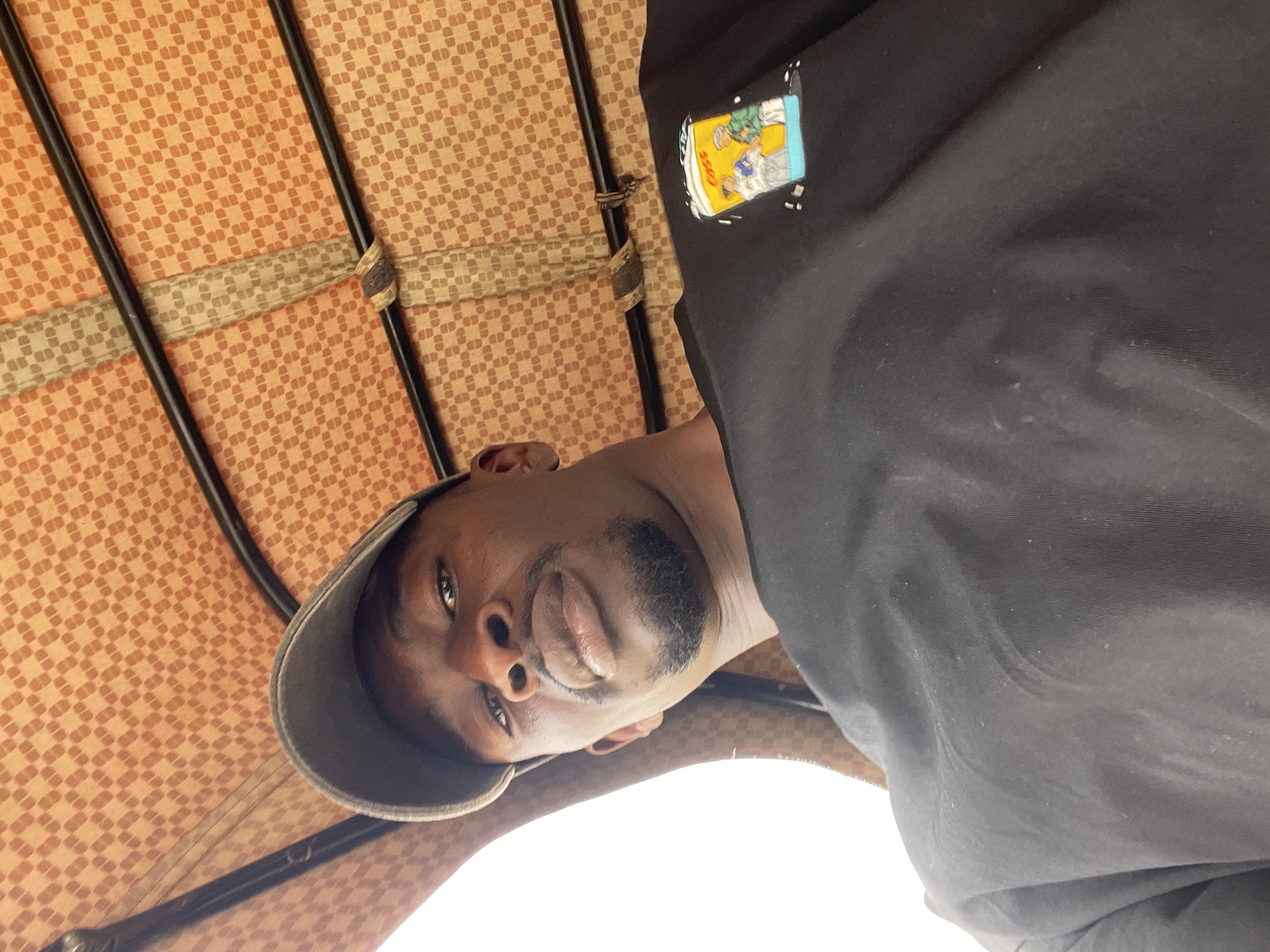 The image shows a man looking down at the camera. He has dark skin, a beard, and is wearing a grey cap and a black shirt with a graphic print on the chest. The background appears to be the interior of a vehicle, possibly an auto-rickshaw or similar transport. The interior has an orange checkered pattern with metal frames. Based on the prompt, the location is Jos, Nigeria.
