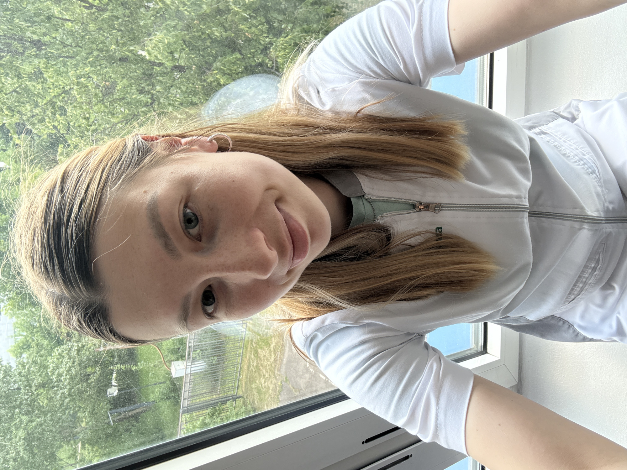 A young woman with blonde hair is taking a selfie. She is wearing a white zip-up jacket over a light green shirt. Her left eye is visible, and she has a slight smile on her face. The background appears to be a window with trees and a fence visible outside. The image seems to be taken indoors, with natural light coming through the window. Based on the distinct architecture of the window frame, which is characteristic of Stalin-era buildings, the location is likely in Moscow, Russia.