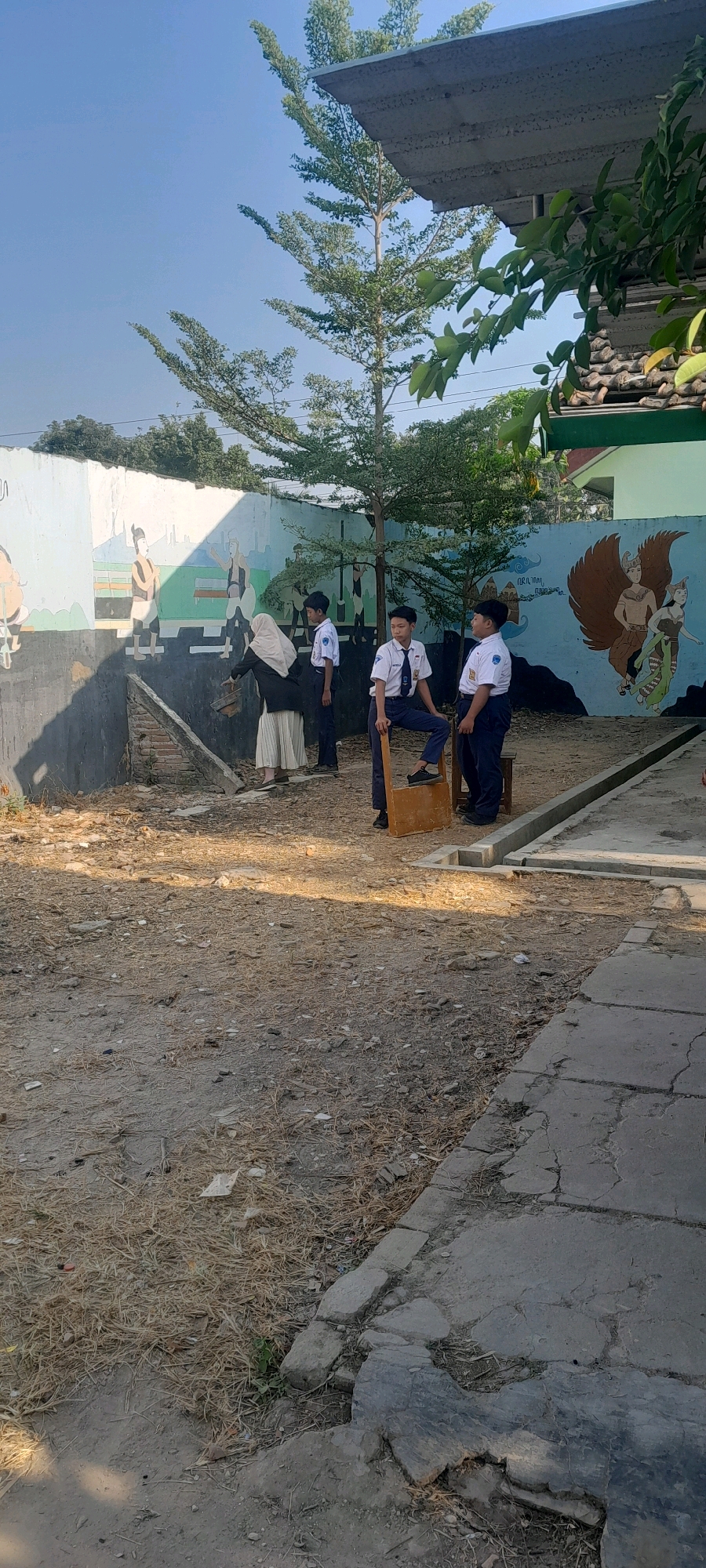 In Tahunan, Indonesia, a group of students in school uniforms are gathered outdoors. The scene is set against a mural-decorated wall depicting figures and a mythical creature. The ground is dry and covered with sparse vegetation and debris. Sunlight casts shadows across the area, suggesting it is daytime.
One student, wearing a white shirt with a blue tie and dark pants, stands on a wooden stool, looking towards the left. Another student, similarly dressed, is seated on the stool, with one leg crossed over the other, also looking left. A third student, in a white hijab and a long, flowing skirt, is bending down, appearing to interact with the ground or the base of the wall. Two more students in similar school uniforms are standing further back, observing the scene. The wall behind them features a colorful mural with various figures, including a Garuda-like bird on the right side. The environment suggests an outdoor school setting or a public space with artistic decorations.