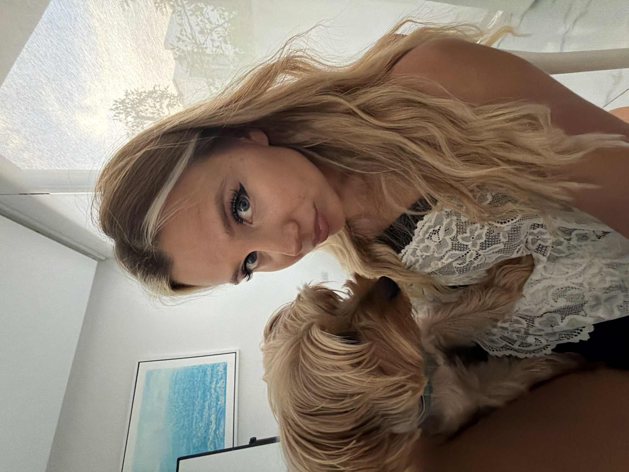 A young woman with long blonde hair is holding a small, tan dog on her left side. She is looking at the camera with her head slightly tilted. The woman is wearing a white lace top over a dark garment. The dog is facing to the left, with its head turned slightly towards the woman. In the background, to the left, is a framed picture of a body of water. To the right, there is a sheer curtain with light coming through it, casting shadows of trees and structures. The overall environment appears to be indoors, possibly a home, with natural light filtering in. The location is specified as Thep Krasatti, Thailand.