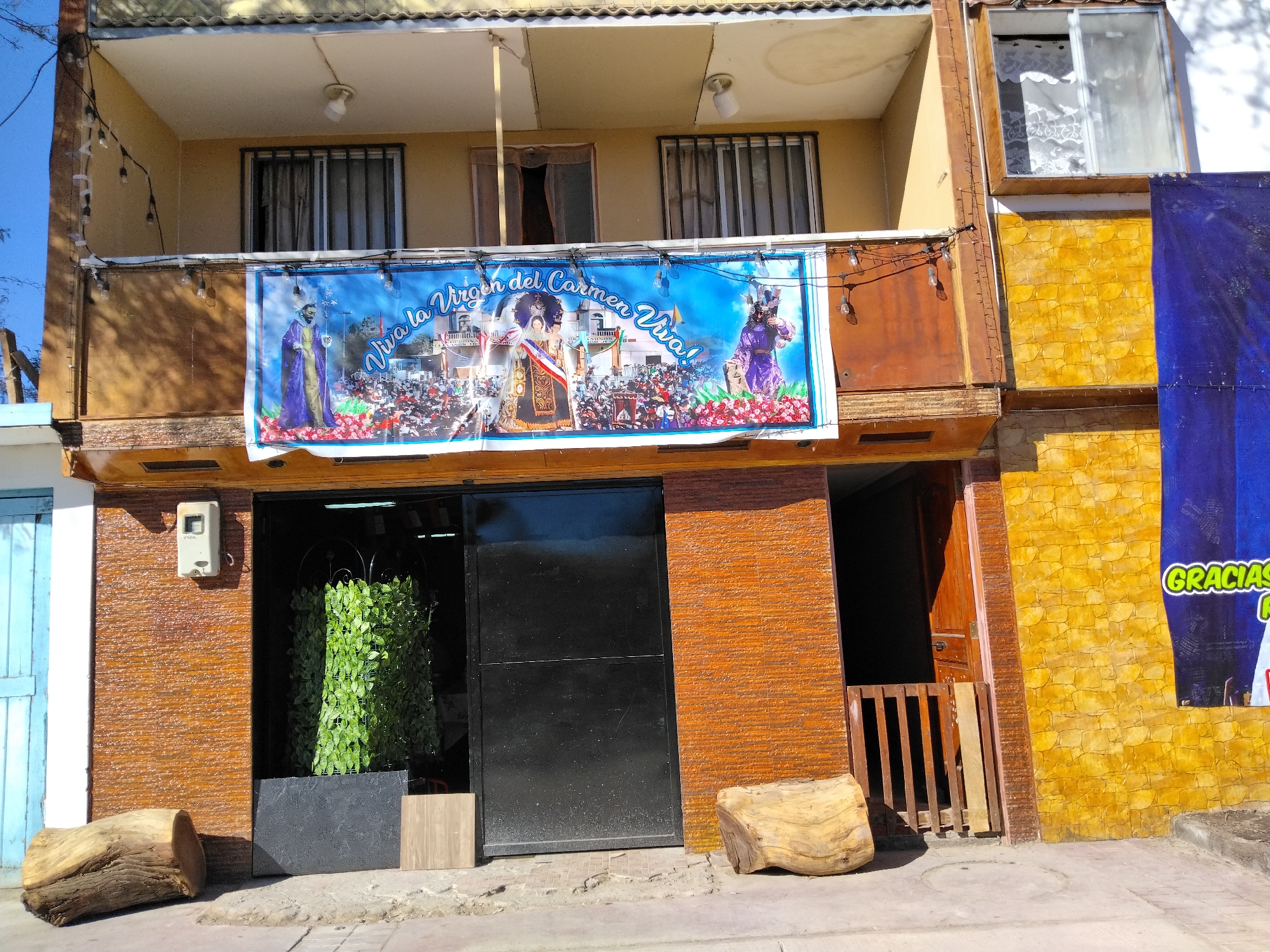 This is a medium shot of a two-story building, likely a business or residence, with a bright blue banner hanging from the second-floor balcony. The building's facade is a mix of textures and colors, with the upper story painted a pale yellow and featuring barred windows. The lower story is clad in rough, reddish-brown brick, with a large black metal door or gate in the center. To the right of the door, a wooden gate blocks access to another entrance.
The banner, which dominates the upper portion of the image, displays a religious scene. It features figures that appear to be saints or religious leaders, surrounded by a crowd of people. The text "Viva la Virgen del Carmen Viva!" is prominently displayed across the banner in a stylized font. Several string lights are draped across the balcony railing, illuminating the banner from above.
The setting appears to be outdoors during the daytime, with clear blue skies and bright sunlight casting shadows. To the left of the building's entrance, a large, rough-hewn wooden log rests on the sidewalk, and to the right, another wooden log of a similar style is positioned near the wall. A dark gray planter with lush green foliage is visible behind the black metal door. On the right side of the building, a large blue banner with yellow text is partially visible, with the word "GRACIAS" legible. The overall scene suggests a celebration or religious event is being honored.