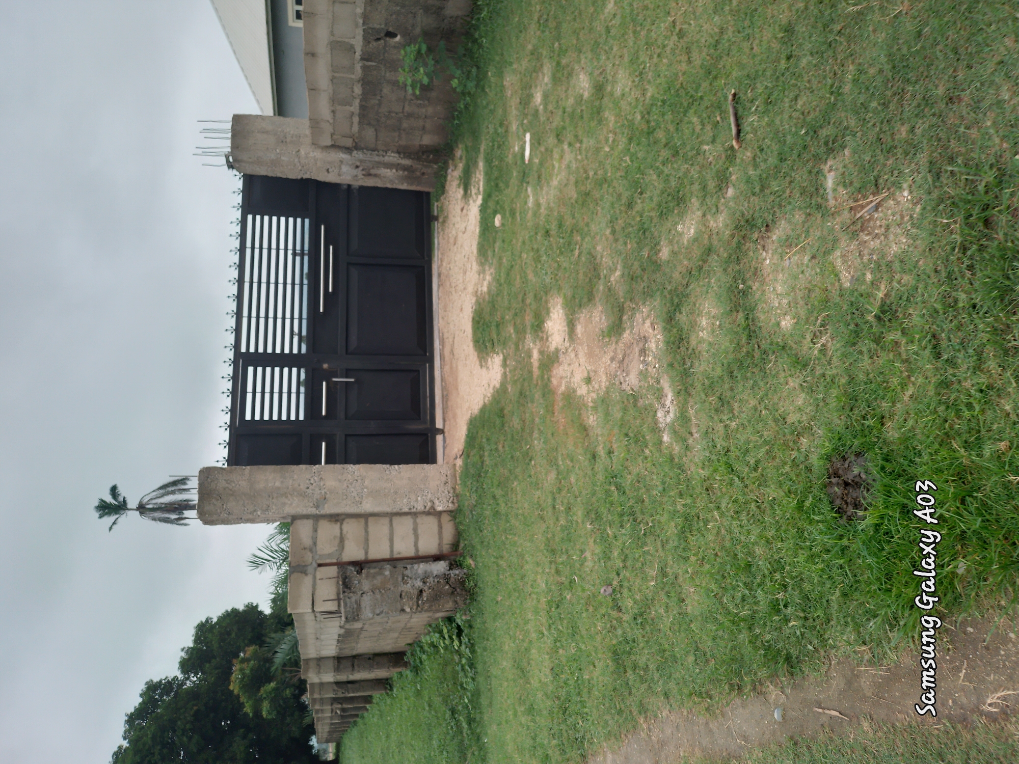 In Doka-Tofa, Nigeria, a black metal gate with horizontal slats stands between two concrete pillars. A wall made of concrete blocks extends to the left of the gate, forming a perimeter. To the right of the gate, a light grey building with a white roof is visible, partially obscured by the gate and wall. The area in front of the gate is a patch of dry earth, transitioning into a grassy yard. The sky is overcast with a light grey cloud cover. A palm tree is visible on top of the left concrete pillar. The bottom of the image displays a watermark reading "Samsung Galaxy A03".