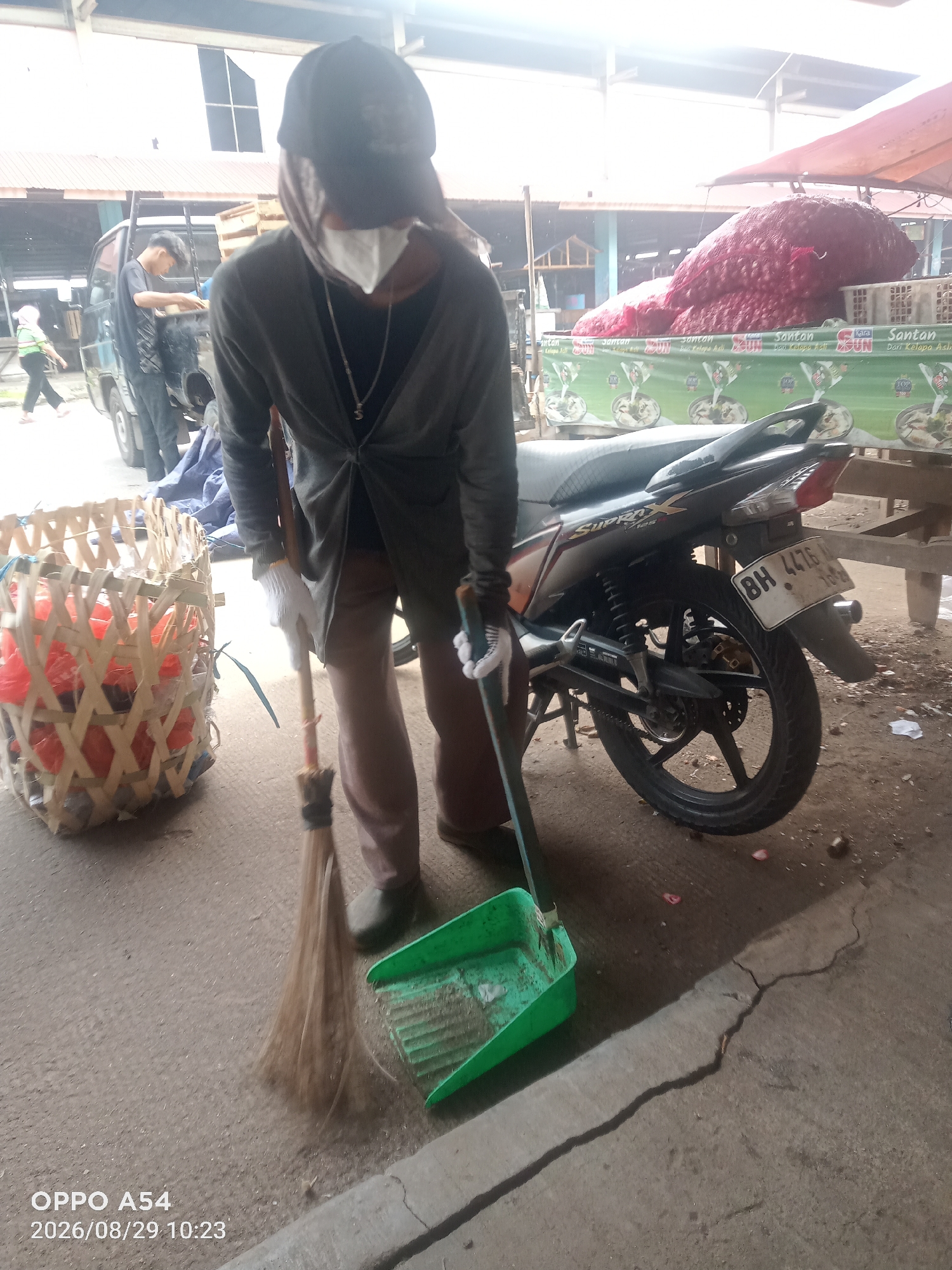 A person wearing a dark baseball cap, a grey cardigan over a dark shirt, brown pants, and white gloves is sweeping the ground with a broom and a green dustpan. The person is also wearing a white mask that covers their nose and mouth. To the left of the person is a woven bamboo basket filled with red plastic bags. Behind the person, a black motorcycle is parked. In the background, a large pile of red onions is visible under a red tarp. There are also other people and vehicles in the distance, suggesting a market or roadside setting. The timestamp at the bottom left indicates the image was taken on August 29, 2026, at 10:23 AM. The location is Jambi, Indonesia.