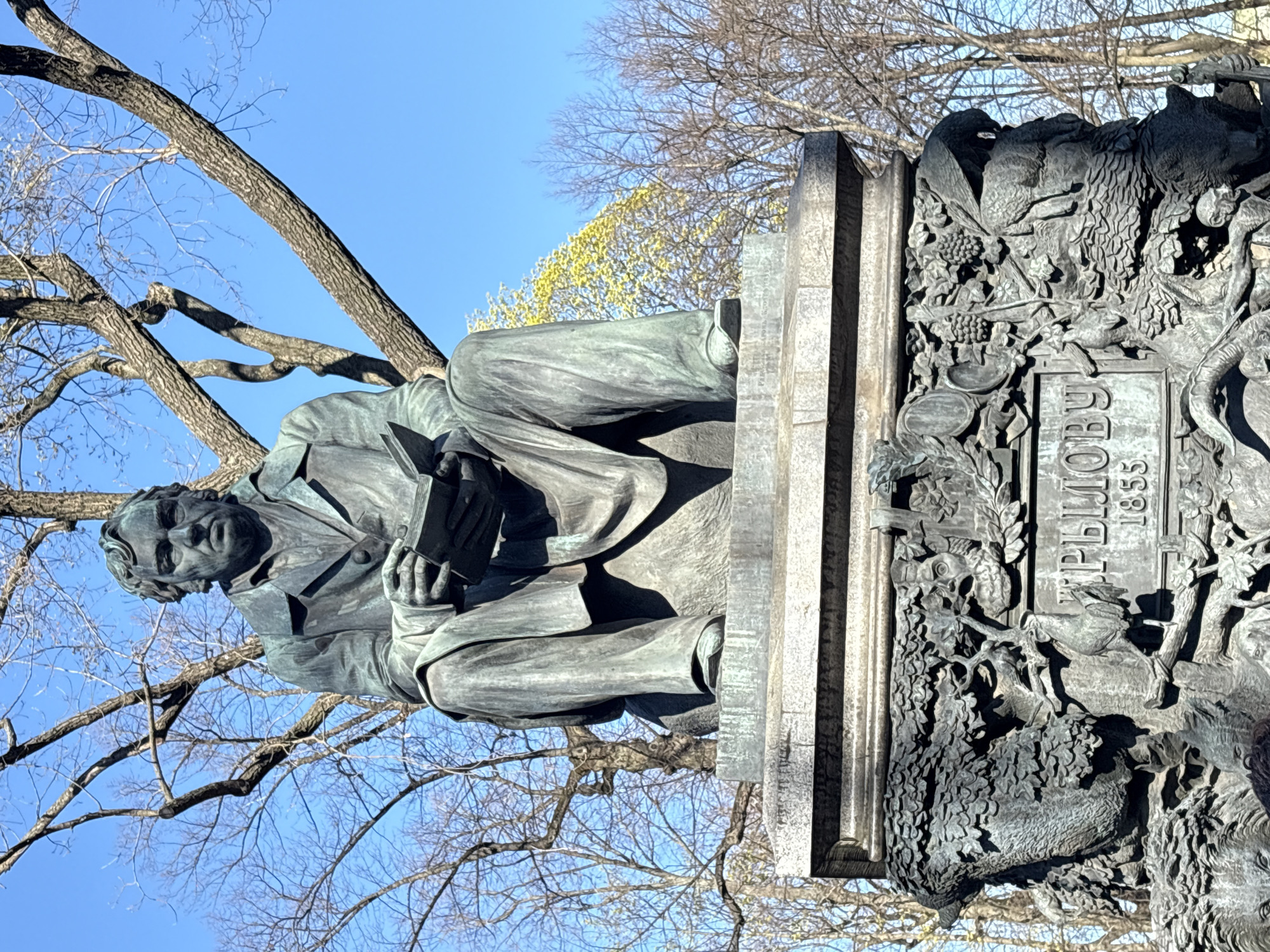 The image shows a monument to Ivan Krylov in Saint Petersburg, Russia. A bronze sculpture of Krylov sits on a stone pedestal, reading a book. The pedestal features intricate carvings of animals, foliage, and a plaque with the inscription "КРЫЛОВУ 1855". Bare tree branches are visible in the background against a blue sky. The monument is located in the Summer Garden.