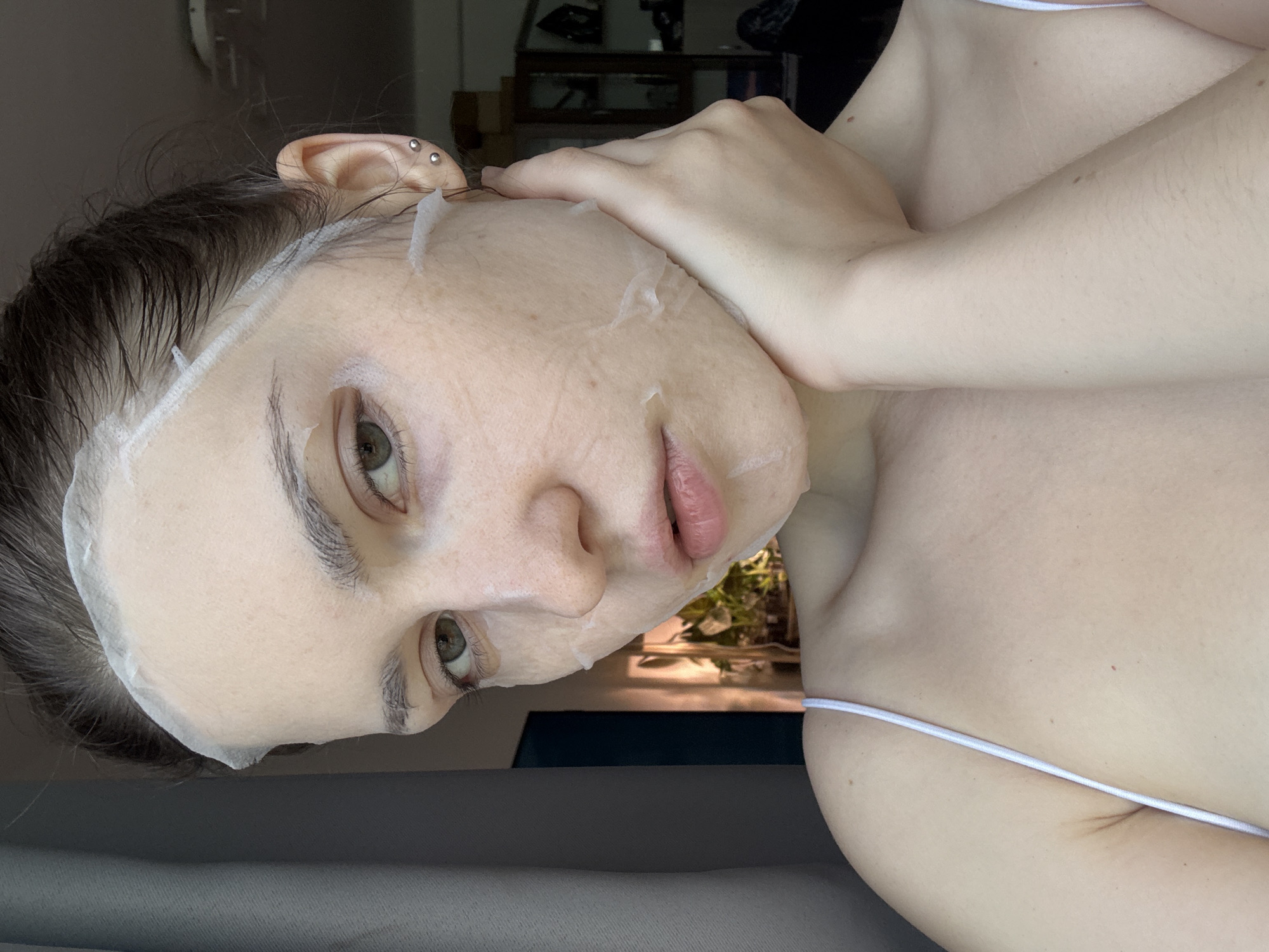 A young woman with fair skin and light brown hair pulled back is seen applying a white sheet mask to her face. The mask covers most of her forehead, cheeks, and nose, with openings for her eyes and mouth. She is wearing a white tank top with a thin strap visible on her left shoulder. Her right hand is gently holding her jawline. Her eyes are green and her lips are slightly parted. The background is softly blurred, showing a window with light filtering through, suggesting an indoor setting. There are hints of furniture and plants in the background. The image appears to have been taken in Saint Petersburg, Russia, though no specific city landmarks are visible.