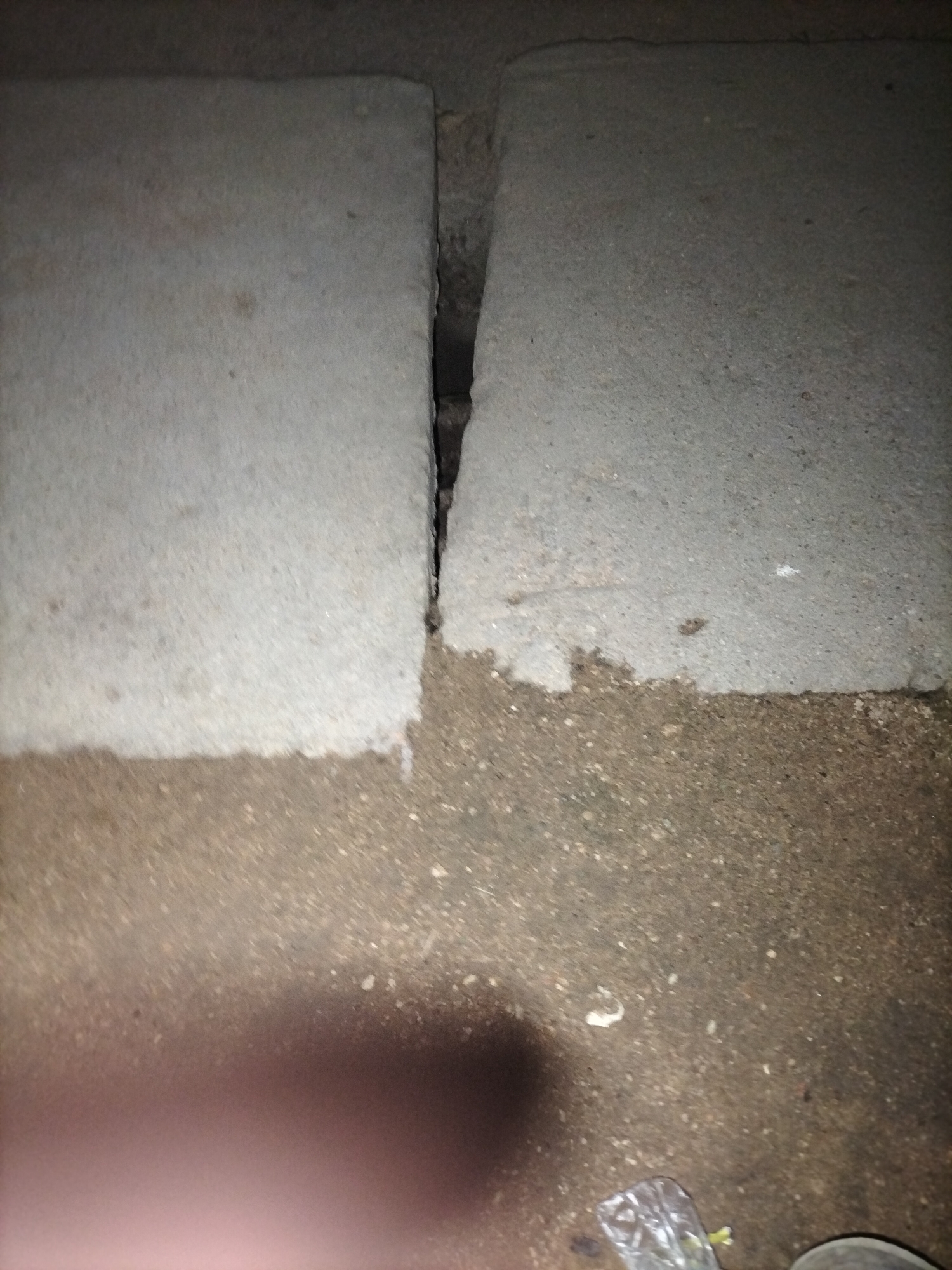 The image shows a close-up, ground-level view of two concrete slabs that have shifted, creating a narrow, dark gap between them. The concrete on the left is lighter in color and appears somewhat worn, while the concrete on the right is darker and has a rougher texture. The ground surrounding the slabs is composed of dirt and small pebbles. In the lower right corner, a clear plastic bag with some green material inside is visible, along with the edge of a dark shoe. A blurred, pinkish shape, possibly a hand or arm, is in the lower left portion of the image. The scene appears to be an outdoor area in Jalingo, Nigeria, possibly a sidewalk or pathway that has experienced ground movement.