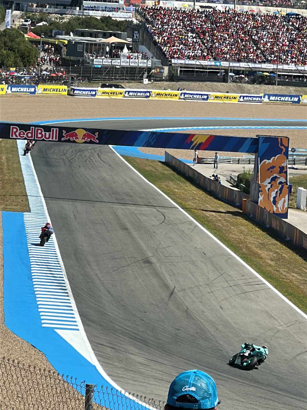 The image shows a section of the Circuito de Jerez-Ángel Nieto racetrack in Jerez de la Frontera, Spain. Crowds of people fill the grandstands in the background, overlooking the track. Banners with "Michelin" are visible along the sides. Two motorcycles are on the track, one in the foreground and another under the "Red Bull" overhead banner. The track has blue and white striped run-off areas. A decorative wall with orange and blue designs borders the track.