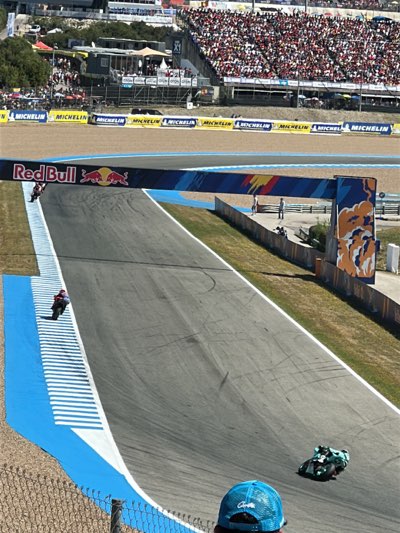 The image shows a section of the Circuito de Jerez-Ángel Nieto racetrack in Jerez de la Frontera, Spain. Crowds of people fill the grandstands in the background, overlooking the track. Banners with "Michelin" are visible along the sides. Two motorcycles are on the track, one in the foreground and another under the "Red Bull" overhead banner. The track has blue and white striped run-off areas. A decorative wall with orange and blue designs borders the track.
