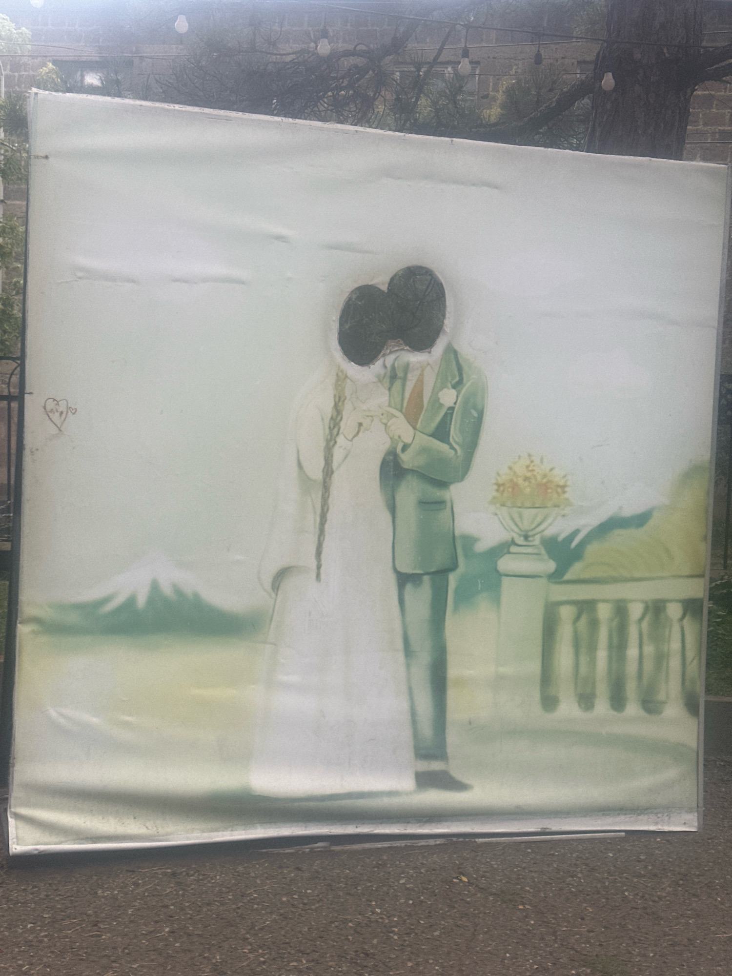 The image depicts a large, rectangular backdrop with two cutouts for faces, designed to resemble a wedding scene. The backdrop is positioned outdoors on a gravel surface, with a tree and some hanging lights visible in the background. The backdrop itself appears to be made of a white material, possibly fabric or vinyl, with a painted illustration. The illustration features a bride in a white dress with braided hair and a groom in a green suit, holding hands. The cutouts are roughly in the shape of heads, revealing the darker background behind them. To the left of the figures, a small drawing of a heart with initials is visible on the backdrop. In the background of the painted scene, there is a mountain resembling Mount Fuji, a potted plant in an urn, and a railing. The backdrop is slightly creased and appears to be weathered. The overall scene suggests an outdoor event or attraction, possibly at a wedding venue. The location is stated to be in Gyumri, Armenia.