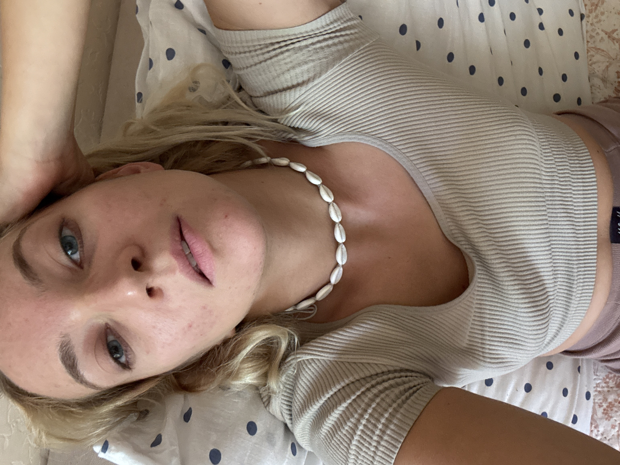 A fair-skinned person with blonde hair and blue eyes is lying on a bed with white and blue polka-dot sheets. The person is wearing a light beige, ribbed, short-sleeved shirt and a white seashell necklace. A small portion of a purple garment is visible. The scene is indoors, likely a private residence. The location is not discernible as Minsk, Belarus, based on visual cues.
