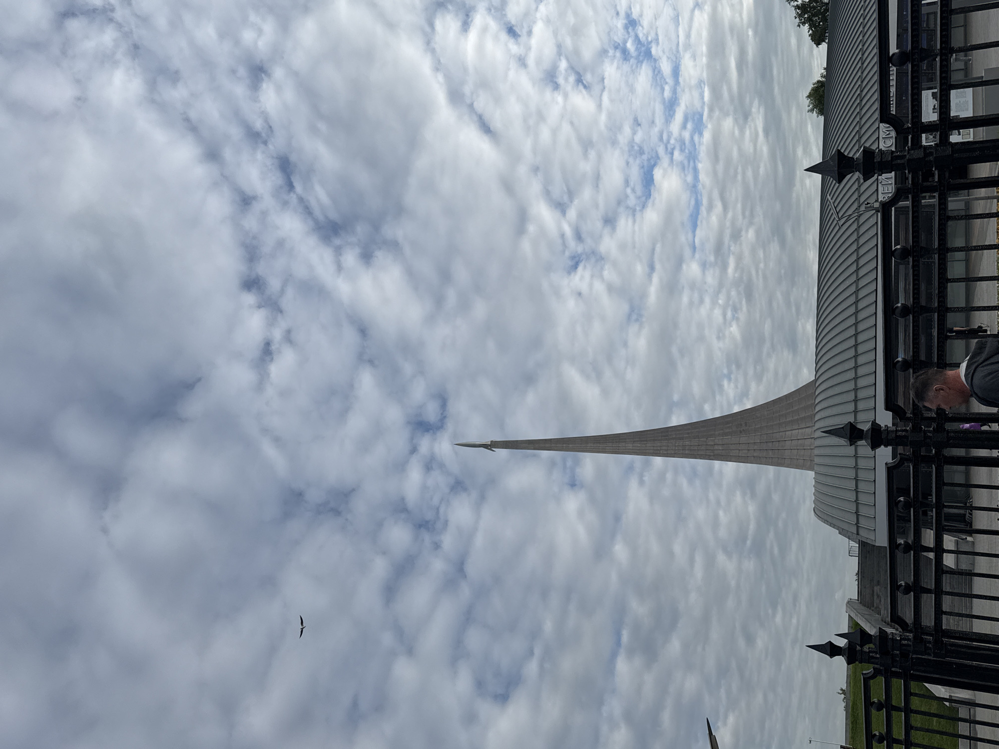 This daytime outdoor shot, taken in Moscow, Russia, features the Monument to the Conquerors of Space and a portion of the Museum of Cosmonautics under a partly cloudy sky. The image is originally rotated 90 degrees clockwise.
The primary subject is the imposing, rocket-shaped Monument to the Conquerors of Space, which tapers dramatically upwards from a wide base. Its surface appears to be a light, ribbed material.
In the mid-ground, a modern building with a distinctive curved, ribbed roof is partially visible, likely part of the Museum of Cosmonautics. A dark, ornate metal fence with spiked finials runs across the lower portion of the frame, separating the viewer from the building. Partially visible Cyrillic text on the building reads "МУЗЕЙ КОСМОНАВТИКИ" (Museum of Cosmonautics).
A person's head and shoulder are visible on the far right, just behind the fence. In the upper left portion of the sky, a dark bird with outstretched wings is seen in flight. The sky itself is dominated by numerous white and grey cumulus clouds, with intermittent patches of blue sky peeking through. The overall scene conveys a sense of grandeur and historical significance.