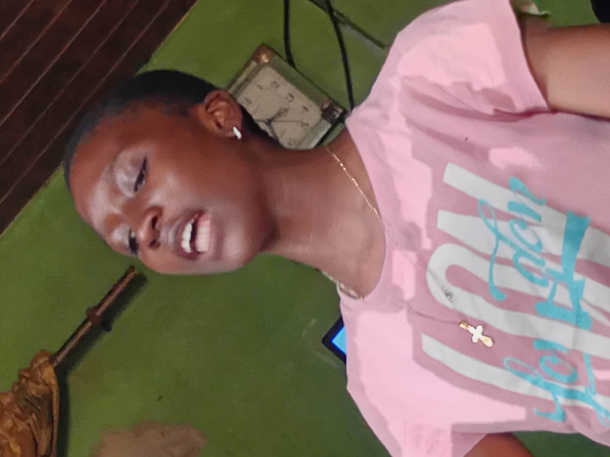 A close-up shot features a young Black person wearing a pink t-shirt with white and teal lettering. The person is looking down and to the left, with their mouth slightly open as if speaking. A thin gold necklace with a cross pendant is visible on their chest. In the background, a clock with Roman numerals is mounted on a green wall. To the left of the clock, there's a wooden object with some brown fabric draped over it, possibly part of a chair or furniture. The overall lighting suggests an indoor setting, possibly during the day. The image is oriented sideways. The text on the t-shirt appears to be a stylized word, possibly "LOVE" in teal and white.