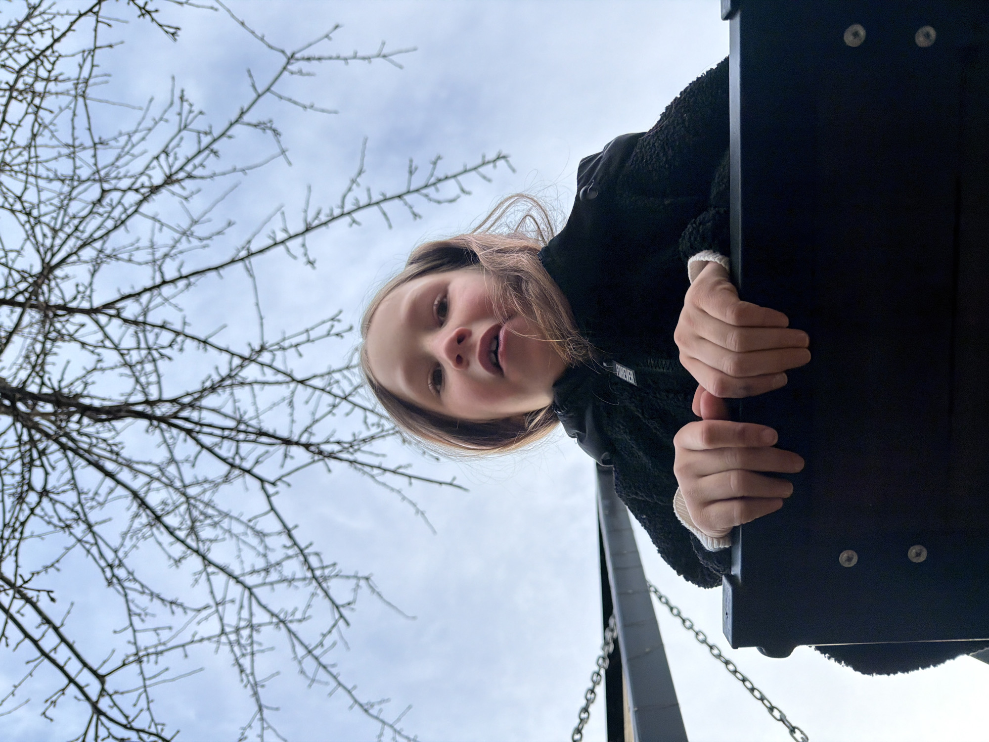 An upward-facing, low-angle photograph captures a young girl looking directly into the camera from a swing in Markovo, Russia. The girl, with light brown hair, is positioned in the lower-right portion of the frame, wearing a dark, possibly fuzzy or sherpa-lined jacket. The word "FOREVER" is visible on a small patch on her jacket near her shoulder. Her hands grasp a dark, rectangular swing seat, from which metal chains are visible descending. The sky above is bright and overcast, indicating daytime. Numerous bare tree branches, possibly coated in ice or hoarfrost, spread across the left and upper sections of the image, suggesting cold weather, likely winter or early spring. The girl's expression appears playful and curious, engaged with the camera.
