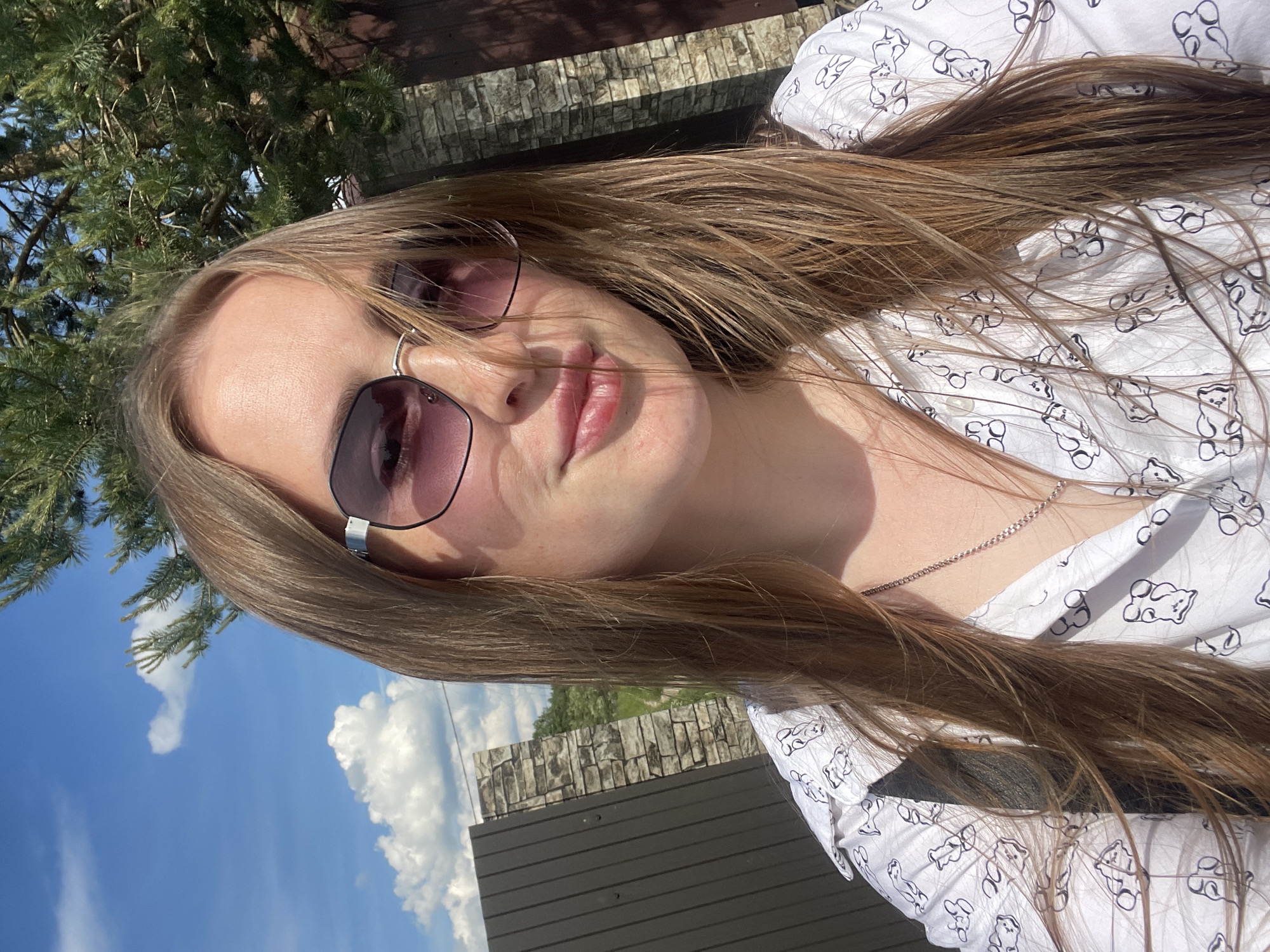 A young adult female is centrally positioned in an outdoor, sunlit portrait taken in Сырково, Russia. She possesses long, straight, light brown hair, which is partially windswept across her face. She wears hexagonal-framed sunglasses with dark, purplish-tinted lenses that reflect the blue sky and green foliage. A subtle smile is evident on her closed lips. Her attire includes a white collared shirt patterned with multiple black outline illustrations resembling gummy bears. A thin silver-colored chain necklace is visible around her neck.
The background displays a clear blue sky with scattered white cumulus clouds. In the upper right quadrant, a pine tree with green needles and cones is prominently featured. To the left, a dark brown, vertically paneled gate or door stands beside a stone pillar constructed from irregular, light-colored stones. Beyond these structures, a dirt path or road and green vegetation suggest a semi-rural environment. A dark brown wooden fence is partially visible to the right. The scene is illuminated by strong direct sunlight, creating distinct highlights and shadows.