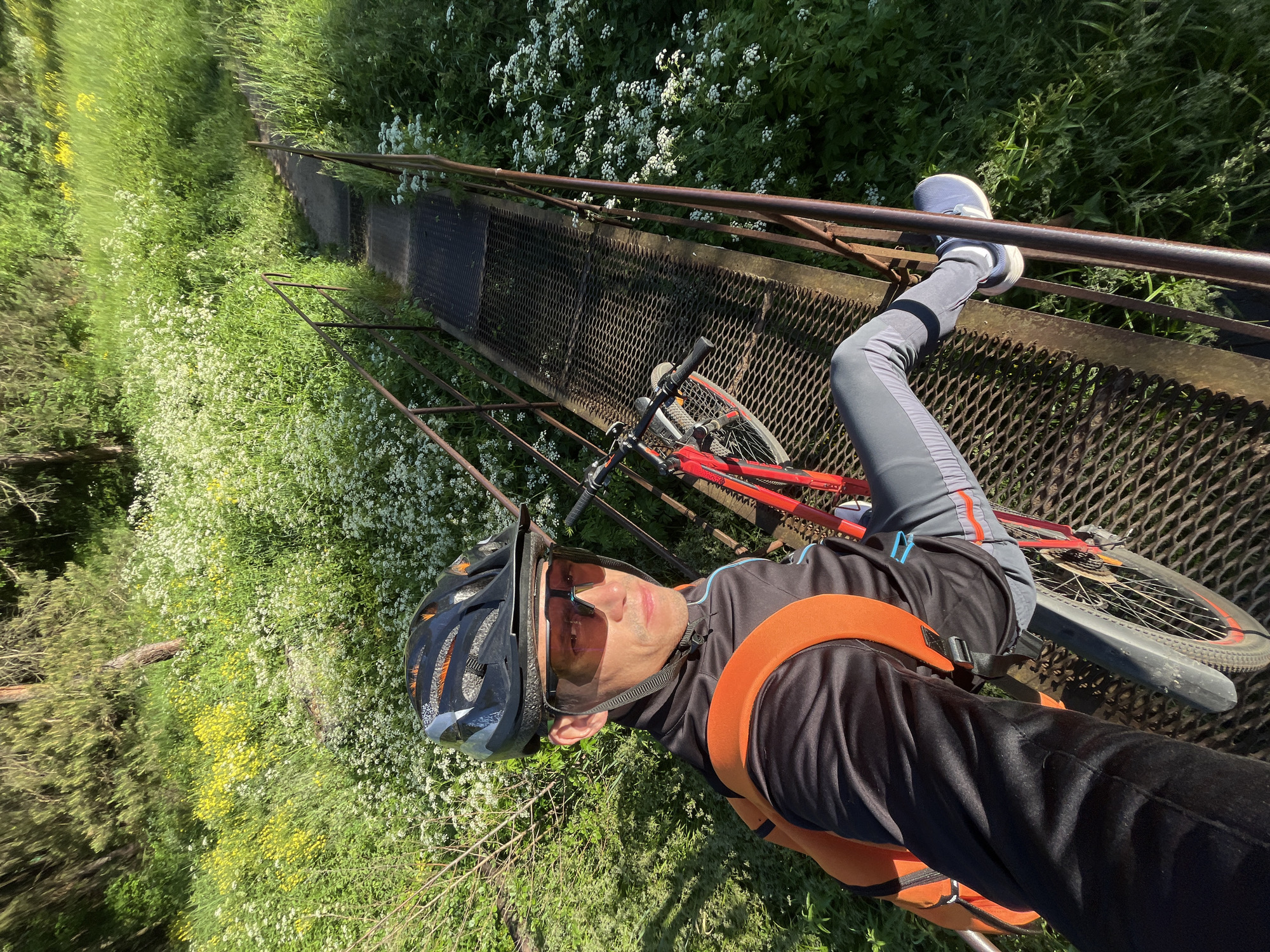 A man wearing a helmet, sunglasses, a black jacket with an orange backpack, and grey pants is taking a selfie while sitting on a metal grate bridge. He is on his bicycle, which is red and black. The bridge is surrounded by lush green grass and white wildflowers. The man and his bike are positioned on the bridge, with the man looking directly at the camera. The scene appears to be outdoors, possibly in a natural setting. The location is identified as Chaikovsky, Russia.