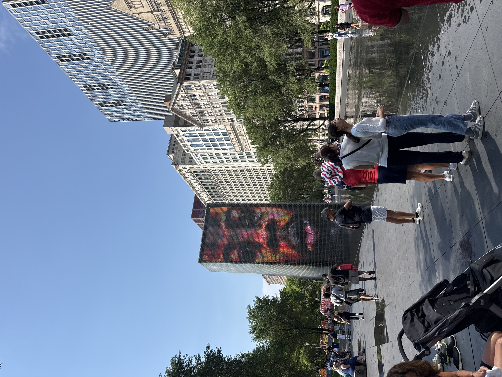 The image captures a scene at the Crown Fountain in Chicago's Millennium Park on a sunny day. The fountain's iconic glass towers display a large, pixelated image of a person's face. A body of water is present in front of the towers, with several people standing on the paved walkway surrounding it. Some individuals are walking, while others are observing the fountain or the surrounding park. Trees and tall buildings, characteristic of the Chicago skyline, are visible in the background. The sky is clear and blue.