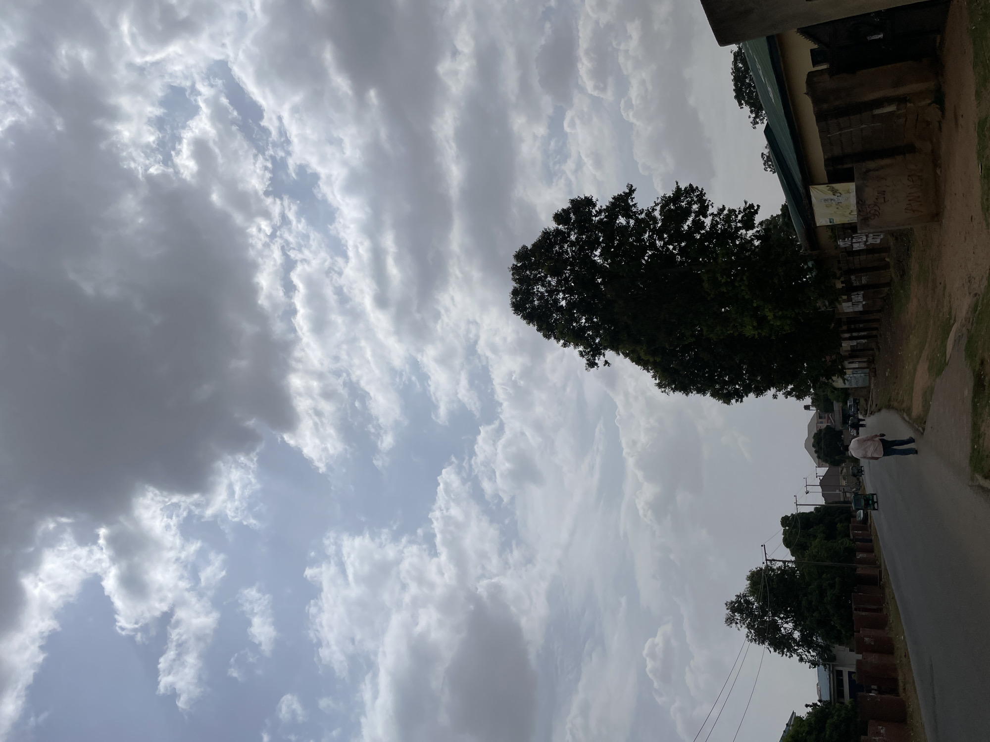 The image captures a street scene in Jos, Nigeria, under a sky filled with dramatic clouds. A large tree dominates the upper-right, while additional trees line the street. A single person walks along the road in the distance. The presence of buildings and utility poles provide context for the urban environment.