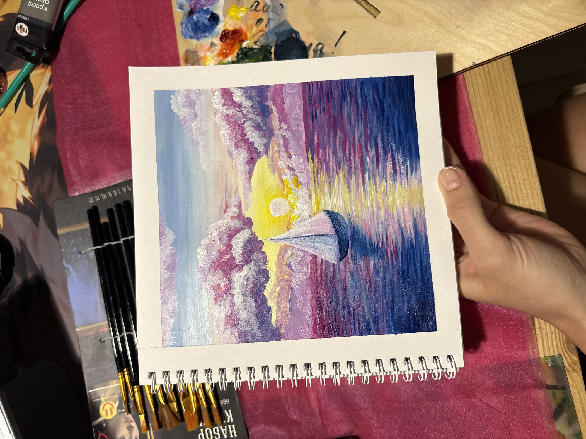The image displays a close-up, high-angle shot of a vibrant, painted sunset or sunrise scene held in a white spiral-bound sketchbook. The location is stated to be in Perm, Russia.
**Subjects and Actions:**
A light-skinned human hand, with a visible thumb resting on the bottom white border, holds the open sketchbook. The painting depicts a serene marine landscape: a bright yellow sun, partially obscured by fluffy, textured clouds, sets or rises over water. The clouds are rendered in shades of pink, purple, and white, contrasting with the lighter blue sky at the top. A single sailboat with a white sail and a dark blue or purple hull is silhouetted against the bright light, positioned slightly to the right of the sun. The water reflects the vibrant colors of the sky and sun, displaying streaks of yellow, pink, purple, and various shades of blue, with darker blues towards the bottom foreground of the painting. The artistic style features distinct, expressive brushstrokes, typical of acrylic or oil paint.
**Objects and Environment:**
The sketchbook itself has a white wire spiral binding on its left edge. Surrounding the sketchbook on a wooden surface, various art supplies are visible. To the left, a retail package of art brushes, featuring golden ferrules and black handles, is partly visible. Russian text "НАБОР КИ" (Set of Brushes) and a yellow smiley face logo are discernible on the packaging. A ruler-like scale with numbers (5, 6, 7, 8, 9, 10, 11, 12) is also part of this packaging. To the right, a green tube of "ГАММА" (Gama) brand paint (a Russian brand) with the word "КРАСКА" (paint) in Russian and the number "0040" is visible, along with a green pencil. Adjacent to the paint tube is an artist's palette, bearing multiple distinct blobs of paint in colors such as red, blue, purple, yellow, and orange, indicating recent use. The immediate surface beneath the art supplies and to the right is covered by a patterned fabric featuring stylized brown and orange shapes. Below this fabric and the sketchbook, a textured, magenta-pink fabric or mat is visible, covering a wooden surface, likely a desk or table. In the far top-left, a dark, featureless object or surface is present.