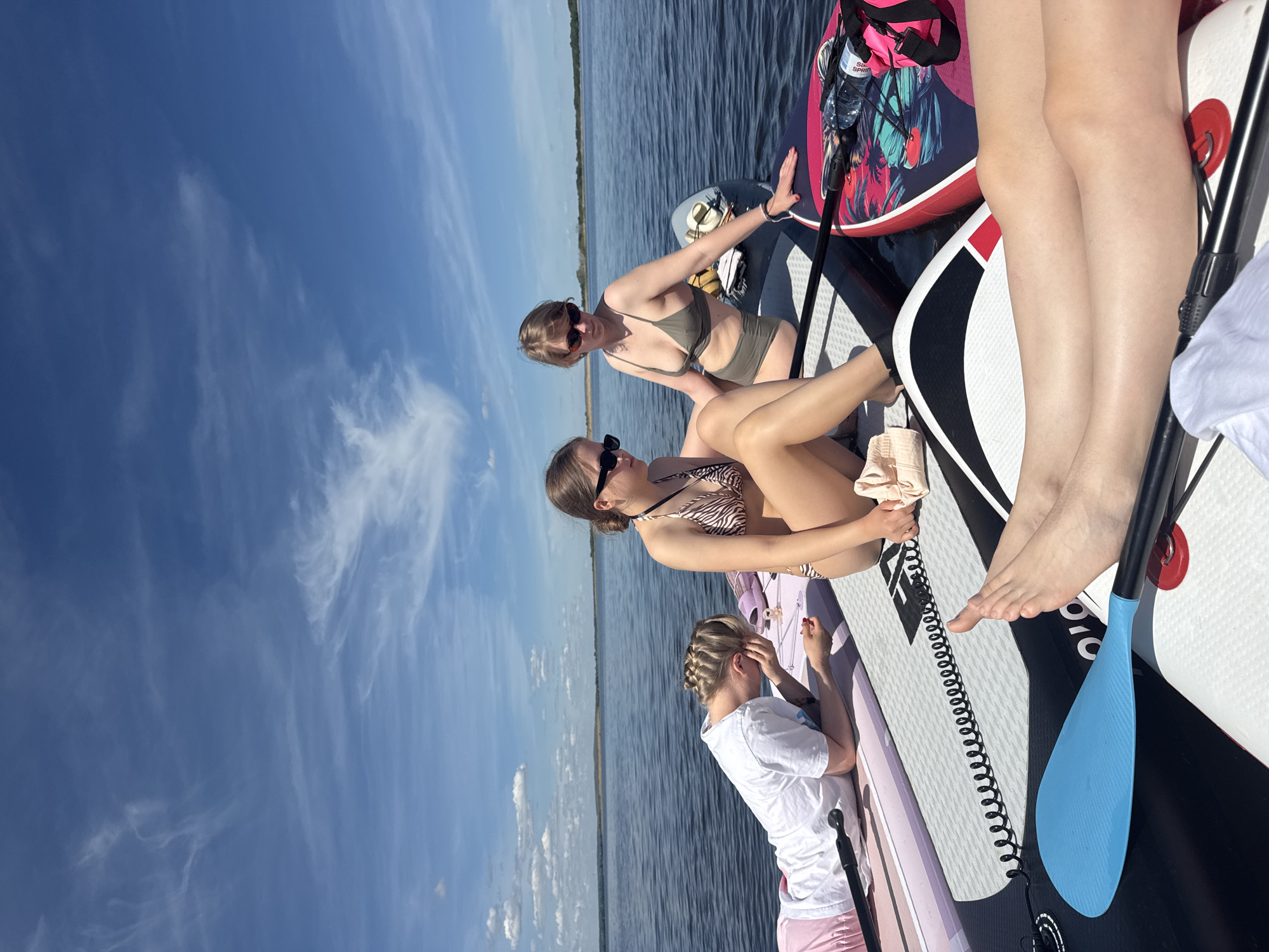 Three young women are depicted relaxing on paddleboards on a large body of water under a clear blue sky with some wispy clouds. The scene is set in what appears to be a lake or wide river, consistent with the location context of Усть-Курдюм, Russia, on the Volga River. A distant, low-lying shoreline is visible on the horizon.
Two of the women are dressed in bikinis and sunglasses, while the third wears a white t-shirt and light-colored shorts, her hair styled in braids. They are positioned across multiple paddleboards, which include designs in black and white, red and black with a tropical pattern, and a light pink/purple. One woman in a dark olive-green bikini sits on a black and white board, reaching towards a red and black board that has a "Sea Sport" branded water bottle attached. Another woman in an animal-print bikini sits on a pink/purple board, holding a light fabric. The third woman rests on her board, appearing contemplative or engaged with something on the board.
Visible objects include several paddleboards, a light blue paddle blade, water bottles, sunglasses, and a coiled black leash. The activity taking place is clearly leisure and sunbathing on the water. The bright sunlight indicates it is daytime, likely a warm, sunny day. The overall mood is relaxed and peaceful.