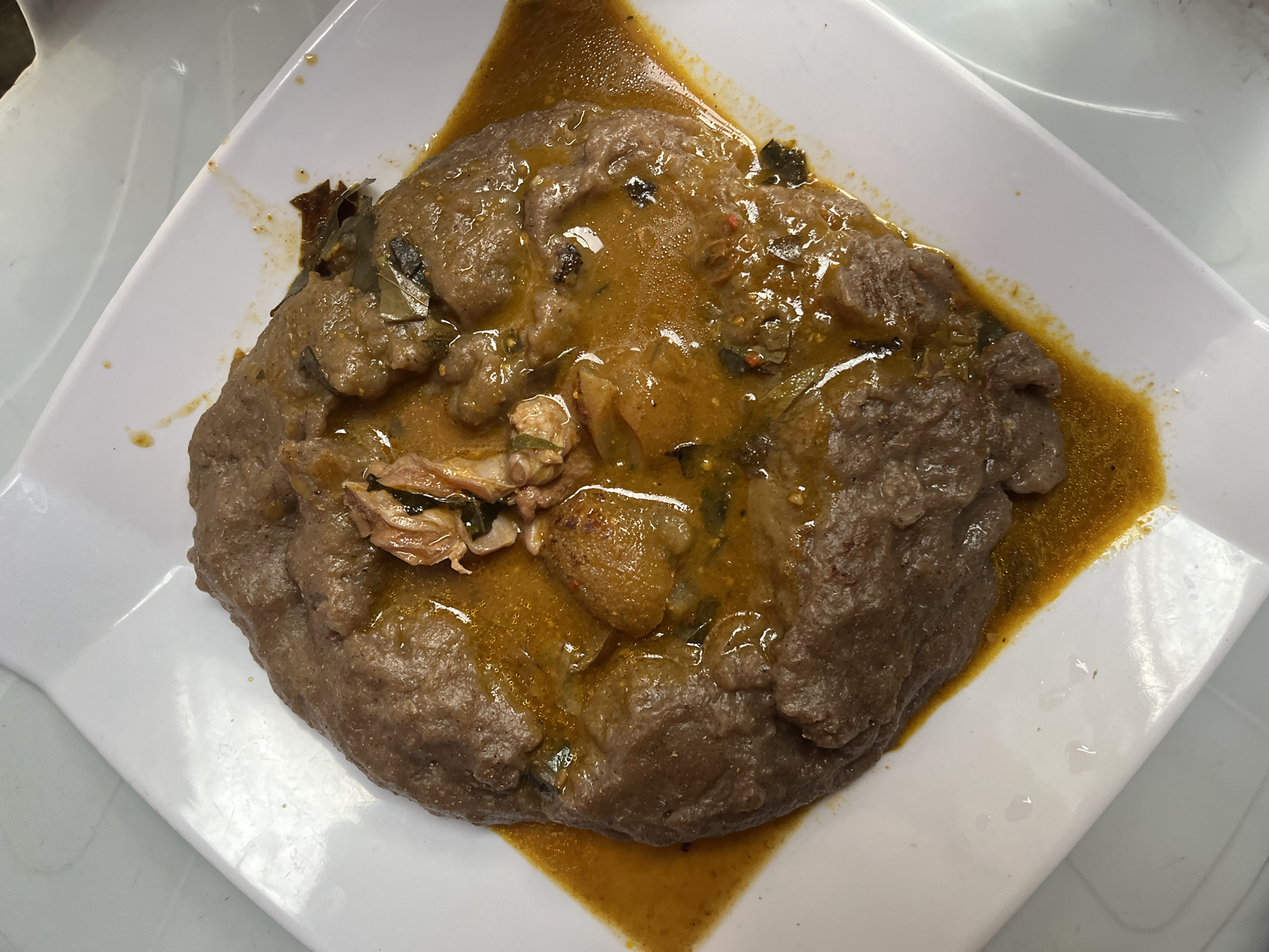 A close-up, top-down shot captures a traditional Nigerian meal served on a white rectangular plate. The central subject is a substantial portion of food, comprising a dark, dense, and somewhat lumpy carbohydrate base, likely a 'swallow' food such as Amala, common in the region. This base is partially covered and generously topped with a rich, glossy, orange-brown stew or soup.
The stew contains various ingredients, including several pieces of cooked protein, which appear to be meat or fish. Some protein pieces are lighter in color and chunky, while others are darker and shredded. Numerous small, dark green specks, indicative of leafy vegetables or herbs, are visible throughout the stew, adding to its texture and visual appeal. The stew's vibrant color suggests the presence of spices and possibly palm oil.
The white plate provides a clean and contrasting background for the earthy tones of the food. The meal appears freshly prepared and ready for consumption. The lighting is bright and even, highlighting the textures and details of the dish. There are no human subjects visible in the frame, and no text is present. The overall scene depicts a hearty and authentic meal, consistent with the specified location context of Iranla, Nigeria.