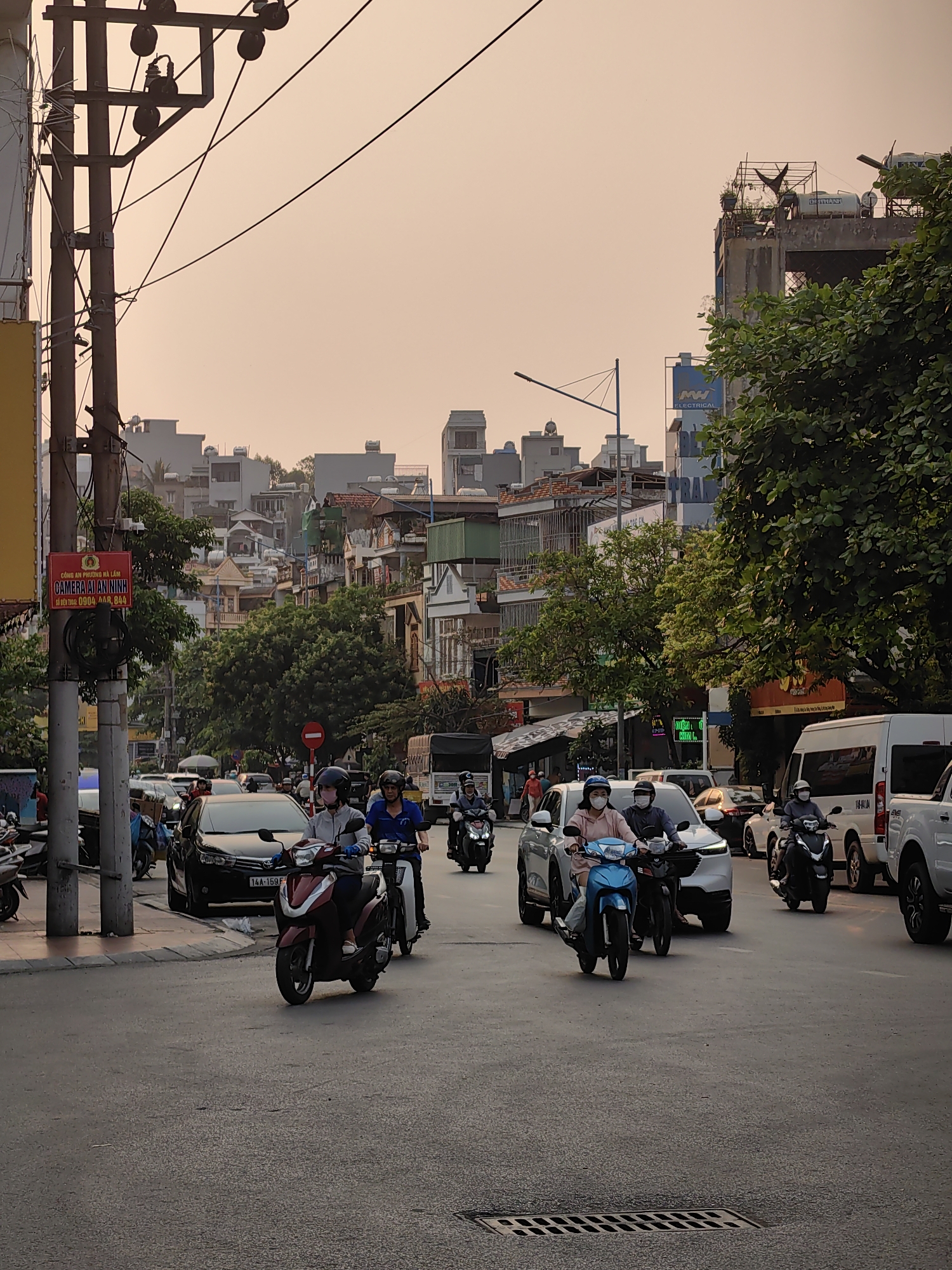 The image shows a street scene in Ha Lam Ward, Vietnam, with moderate traffic composed of cars and motorbikes. Several people are riding motorbikes and wearing masks. There are buildings and trees lining the street. A sign for "Camera AI An Ninh" (AI Security Camera) is visible on a utility pole. The sky is a pale orange, suggesting either dawn or dusk. There is a sign for "MW Electrical" located in the background above the road.