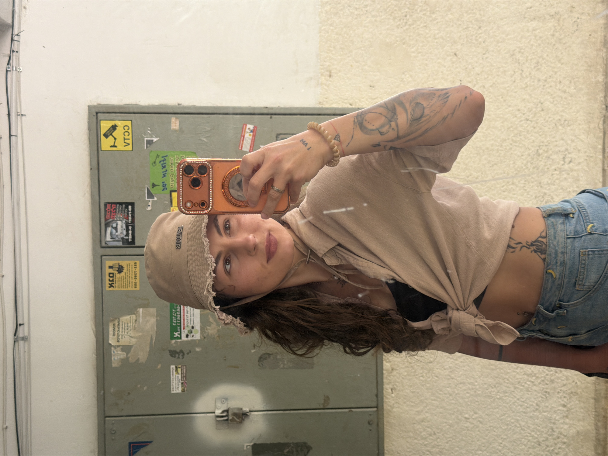 A young woman is captured in a mirror selfie, holding a rose gold-colored smartphone with a sparkling, embellished camera case. She is reflected in what appears to be a large mirror, showcasing her casual attire and several tattoos. She wears a light-colored, possibly beige, bucket hat with textured trim, and a matching collared shirt tied at her midriff, revealing a black top underneath. Blue denim jeans or overalls are visible from her waist down. Her dark, wavy hair cascades over her shoulders. She has a subtle smile and light makeup, with a pinkish hue on her lips.
Notable accessories include a ring on her right ring finger and a light-colored coiled bracelet on her left wrist. Intricate tattoos adorn her left arm, resembling a sleeve, and another tattoo is visible below her left ribcage. A small tattoo on her left wrist reads "MA I".
The background, visible through the mirror, suggests an urban or semi-urban outdoor setting in Kiryat Bialik, Israel. It features a grey utility cabinet or electrical box covered with various stickers. A yellow sticker shows a CCTV camera icon with the text "CCTV". Another yellow sticker has Hebrew text, "הגנה אקטיבית מ 050-938-1500" (Active protection from 050-938-1500). A green sticker reads "גרר לוי 052-259-3323" (Levi Towing 052-259-3323). Other smaller, less legible stickers are also present. The walls surrounding the cabinet are light-colored, one appearing white and another with a rough, textured finish in a beige or light brown tone. Wires are visible running along the top left of the white wall. The scene appears to be set during the daytime, with ample ambient light.