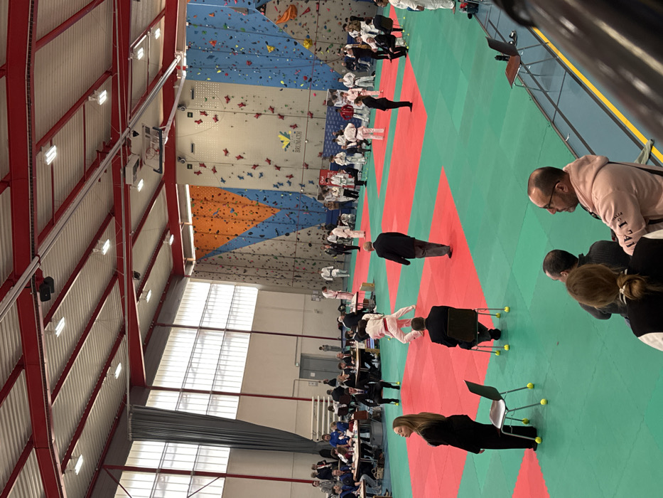 The image displays an indoor sports facility, identified as being in Brumath, France, evidenced by a prominent "BRUMATH" logo on a climbing wall. The venue features a high ceiling with exposed red structural beams and corrugated white roofing, illuminated by natural light from large windows along the left wall and artificial overhead lighting. The right side of the facility is lined with tall, multi-colored climbing walls adorned with numerous handholds. The main floor area is covered with interlocking mats, primarily red and green, delineating various activity zones. Approximately 50-70 individuals are present. Numerous people are dressed in white judo gis, with some wearing colored belts; two individuals in gis, one with a red belt, are actively engaged in a judo stance on a red mat section in the mid-ground. Several men in dark suits are observed, some seated at tables on the far left, others standing or seated on chairs within the mat area, including one chair in the foreground with yellow tennis balls attached to its legs. Spectators are visible, including three individuals in the lower right foreground with their backs to the camera, observing the scene, and a woman in a dark suit standing in the left foreground. Ceiling-mounted speakers and a red "Oui" logo on a banner in the background are also present.