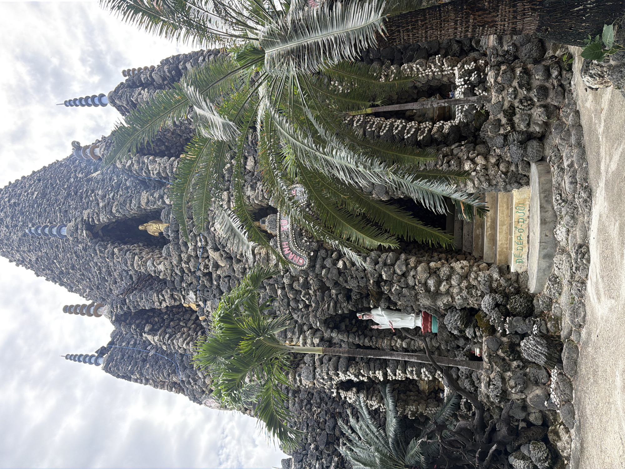 The image captures a unique, ornate structure made predominantly of dark, irregularly shaped stones or coral, resembling a natural grotto or a heavily textured building. Lush green palm trees frame the scene, their fronds extending across the foreground and along the structure, indicating a tropical setting. The sky above is overcast with white and gray clouds.
The structure features multiple spires or pinnacles on its upper levels, some topped with decorative, concentric white discs. Adorning the rock walls are intricate patterns formed by rows of small, white shells, adding a distinct decorative element.
Several figures are visible: a golden statue is nestled within a niche on the upper left side of the structure, while a standing white statue, possibly a religious figure, is positioned closer to the ground on the right. Below the white statue, an entrance or alcove is visible.
To the right of the structure, concrete steps lead up, and a paved path is present. Visible text in green on a concrete surface reads "DE DEP O". Higher up on the rock wall, near the top center, another piece of text, partially obscured and in pink and white, appears to be "CHAMPAL" or a similar word, potentially referencing Cham Pa culture. The scene depicts a static view of this culturally significant and intricately crafted shrine or temple in Cam Ranh, Vietnam.