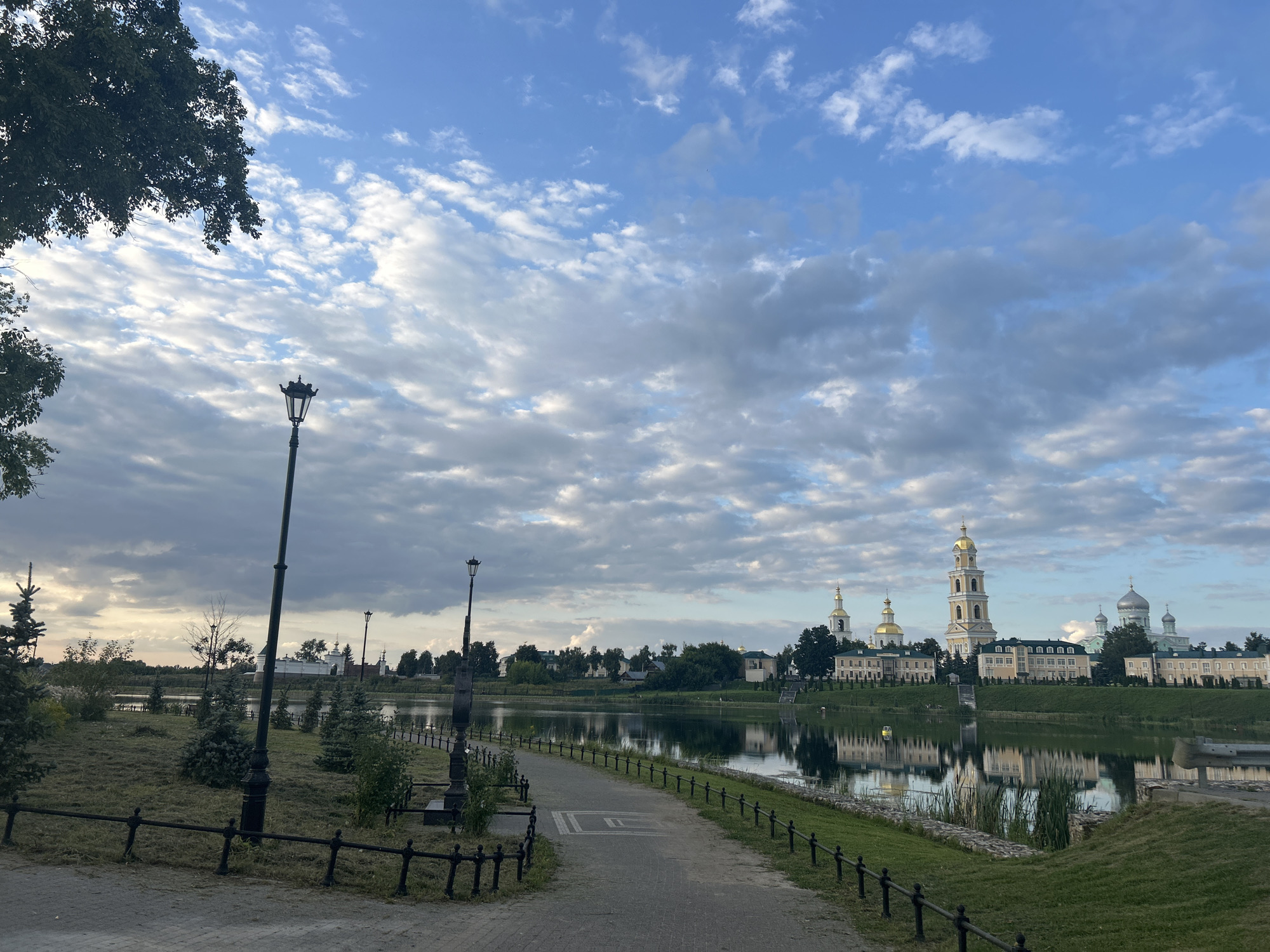 The image depicts a scenic view of the Seraphim-Diveyevo Monastery in Diveyevo, Russia, on a cloudy day. A paved pathway, lined with a black metal fence, curves towards the right, leading the viewer's eye to the monastery complex across a calm body of water. The monastery is characterized by its numerous golden-domed churches and bell towers, with the tallest bell tower prominently featured. The water reflects the sky and the monastery's architecture. On the left side of the pathway, there is a grassy area with scattered trees and decorative shrubs, and a vintage lamppost stands along the path. The sky is a mix of blue and white clouds, with sunlight breaking through in some areas. The overall atmosphere is serene and picturesque.