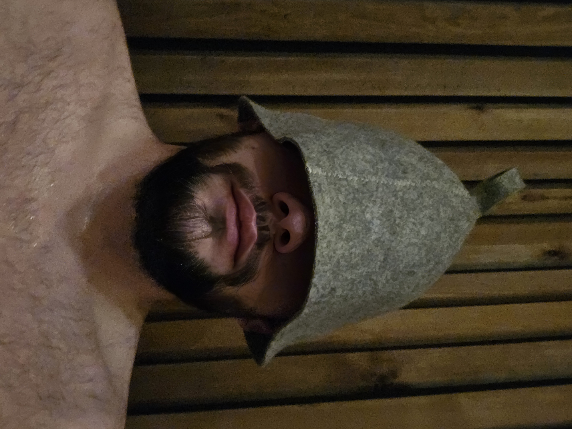 The image presents a close-up, slightly rotated view of a man relaxing in what appears to be a sauna or steam room. The subject is an adult male with a dark beard and visible chest hair, indicating he is shirtless. He wears a traditional grey, conical felt sauna hat, which covers most of his head. His eyes are closed or partially closed, and his mouth is slightly open, conveying a strong sense of relaxation and comfort, possibly from the heat. His skin appears somewhat flushed and moist.
The background is composed of horizontal wooden slats, typical of sauna interiors. The lighting is dim and warm, contributing to the serene and intimate atmosphere. No specific time of day is evident, but the indoor setting with subdued lighting creates a tranquil ambiance. There is no visible text in the image. The overall scene depicts a peaceful moment of unwinding within a hot, therapeutic environment.
