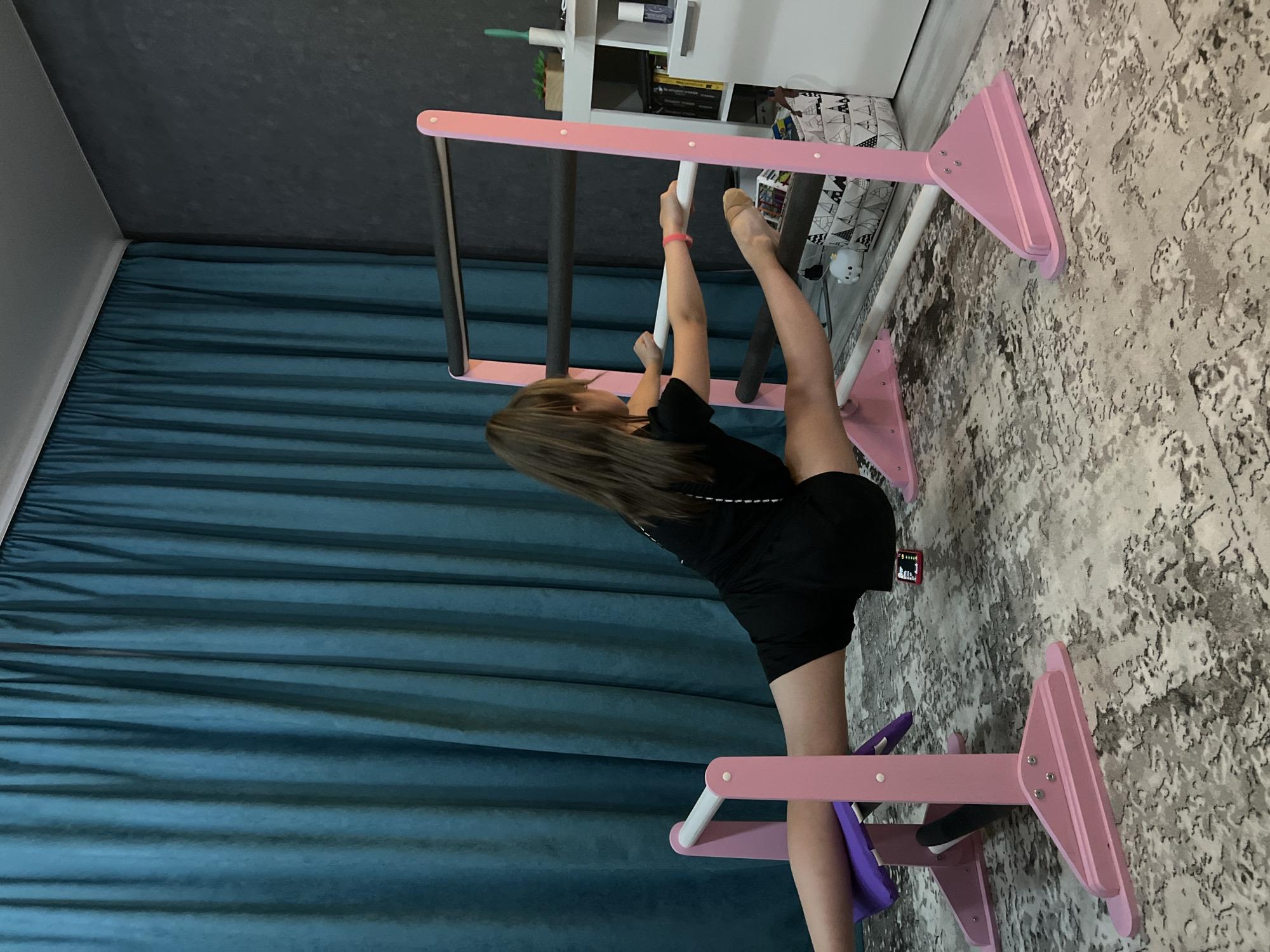 A young girl with long brown hair is performing a split on a pink and white home gymnastics bar set. She is wearing a black t-shirt and black shorts. Her left leg is extended towards the bars, and her right leg is extended behind her. The gymnastics bar set is positioned in a room with a blue curtain and a light gray and white patterned carpet. A white shelving unit is visible in the background. Based on the room decor, it is likely this scene is taking place in a residential setting. It is not possible to confirm the location in the city of Chaikovsky, Russia based solely on the visual information provided.